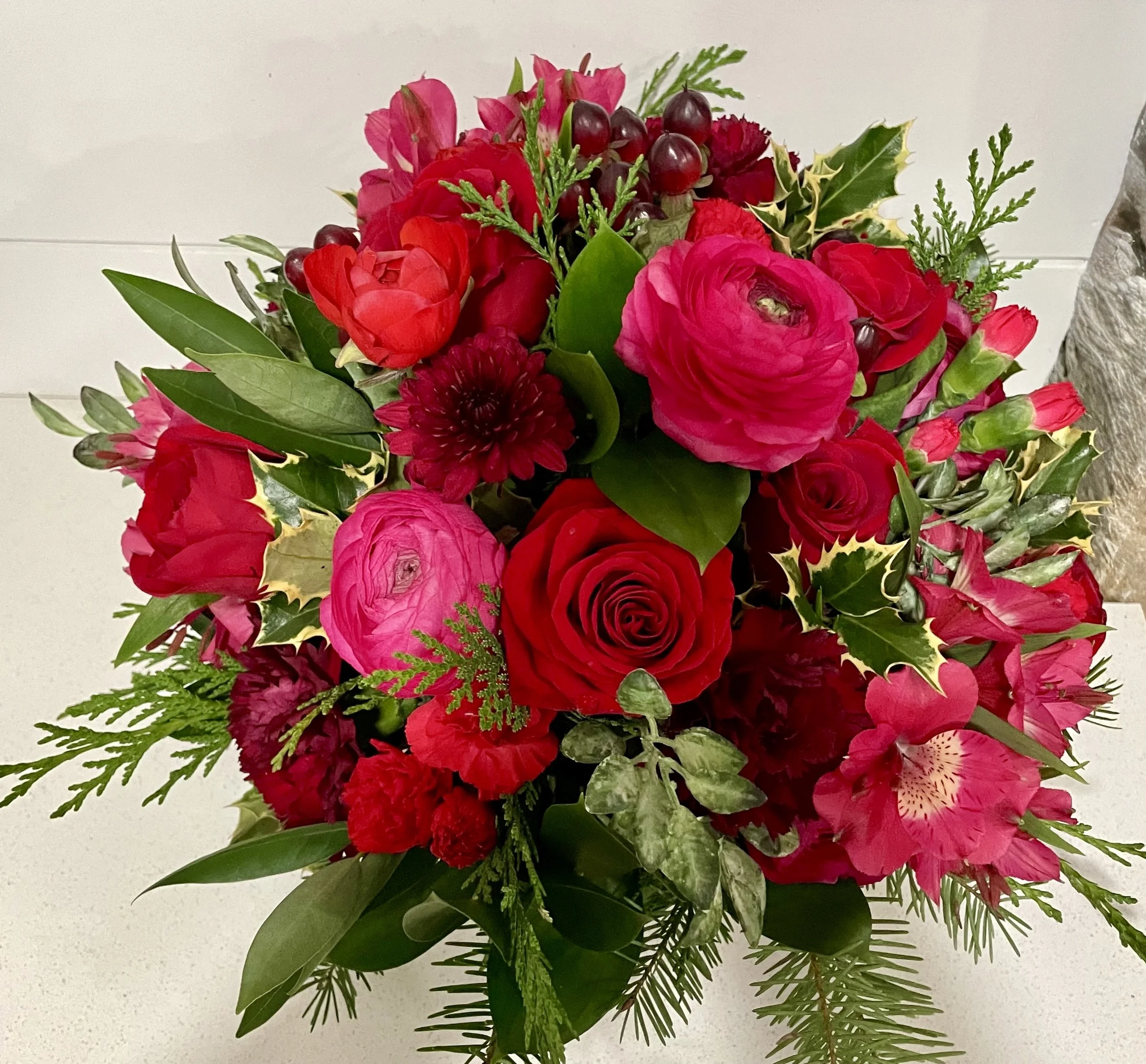 Bouquet of red and pink flowers with green leaves and holly, arranged in a round shape.