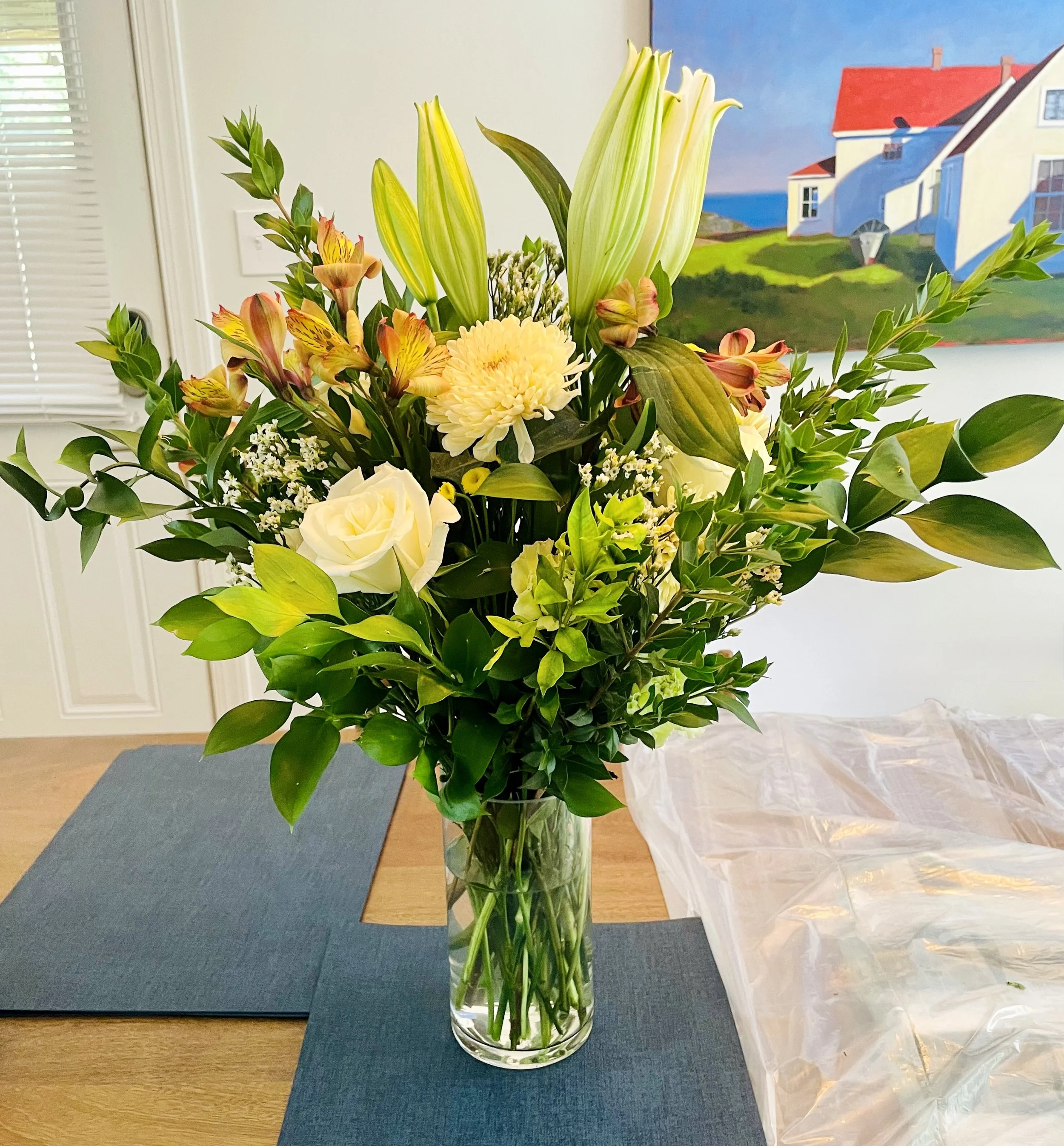 A tall glass vase filled with a large bouquet of white, cream, and orange flowers, including lilies, roses, and chrysanthemums, on a wooden table with blue placemats in a room with a painted landscape on the wall.