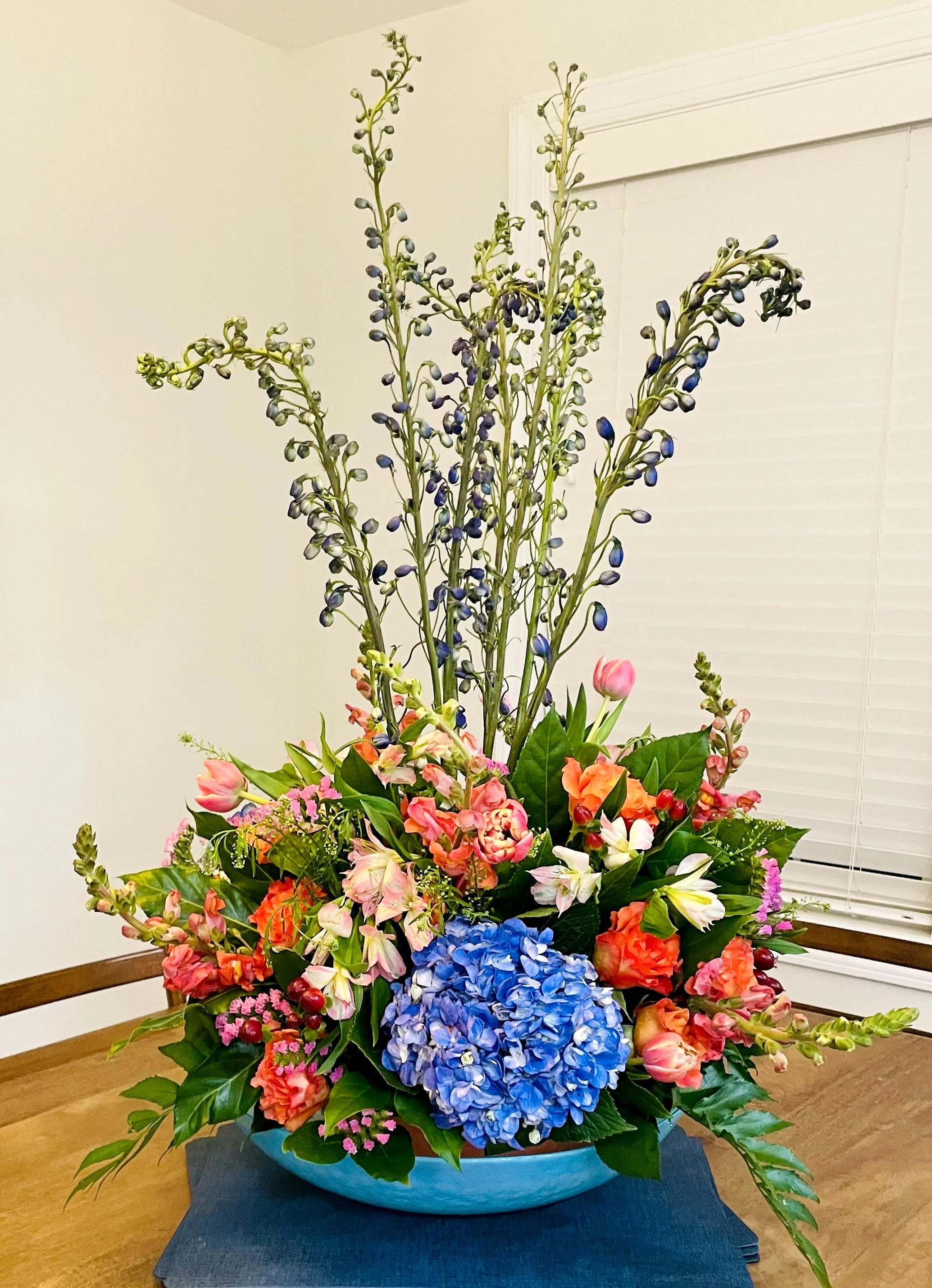 A floral arrangement in a large blue bowl featuring pink, orange, white, and blue flowers with green leaves.