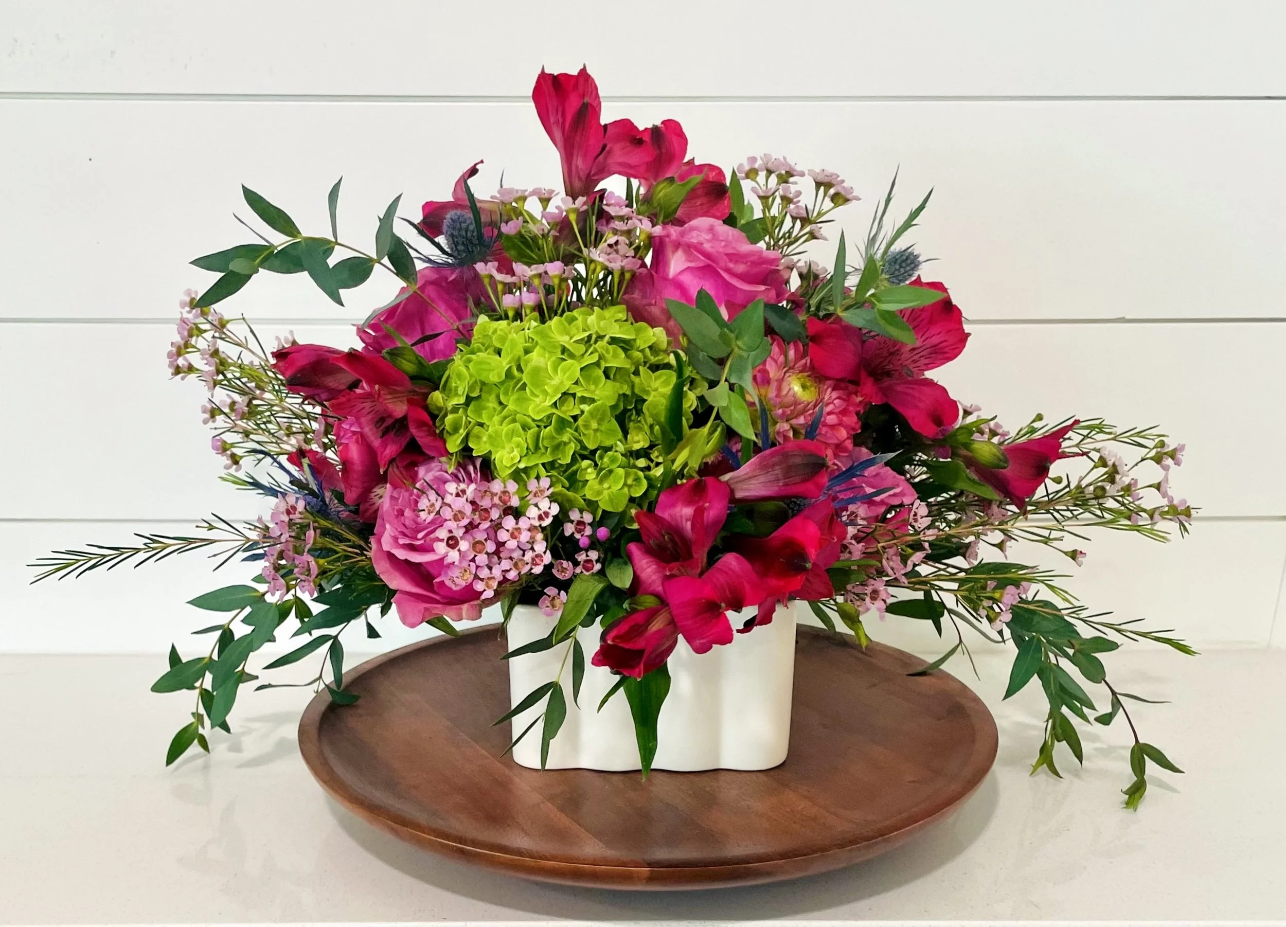 A colorful flower arrangement with pink, red, and green flowers in a white vase, placed on a wooden tray against a white shiplap wall background.