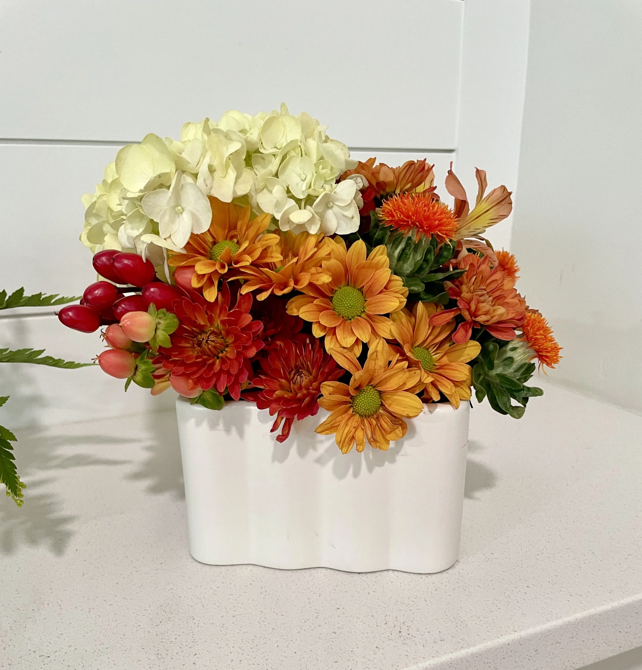 A white ceramic vase with a mix of white, orange, and red flowers, including hydrangeas, daisies, and other blooms, sitting on a white surface against a plain background.