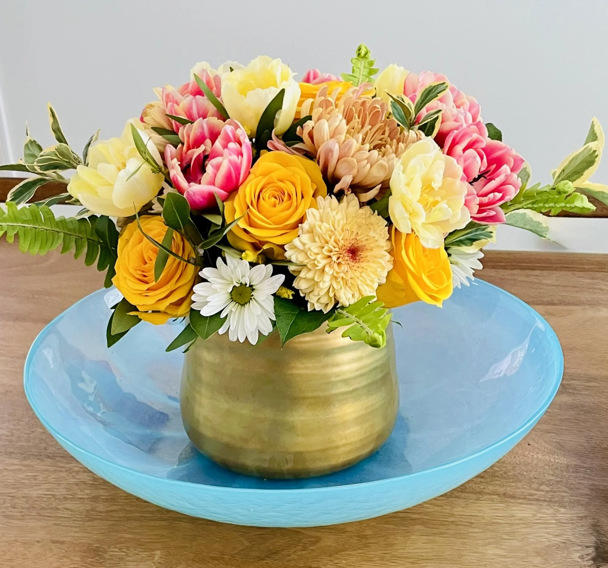 Colorful flower arrangement with yellow roses, pink tulips, white daisies, and beige chrysanthemums in a gold vase on a blue plate.