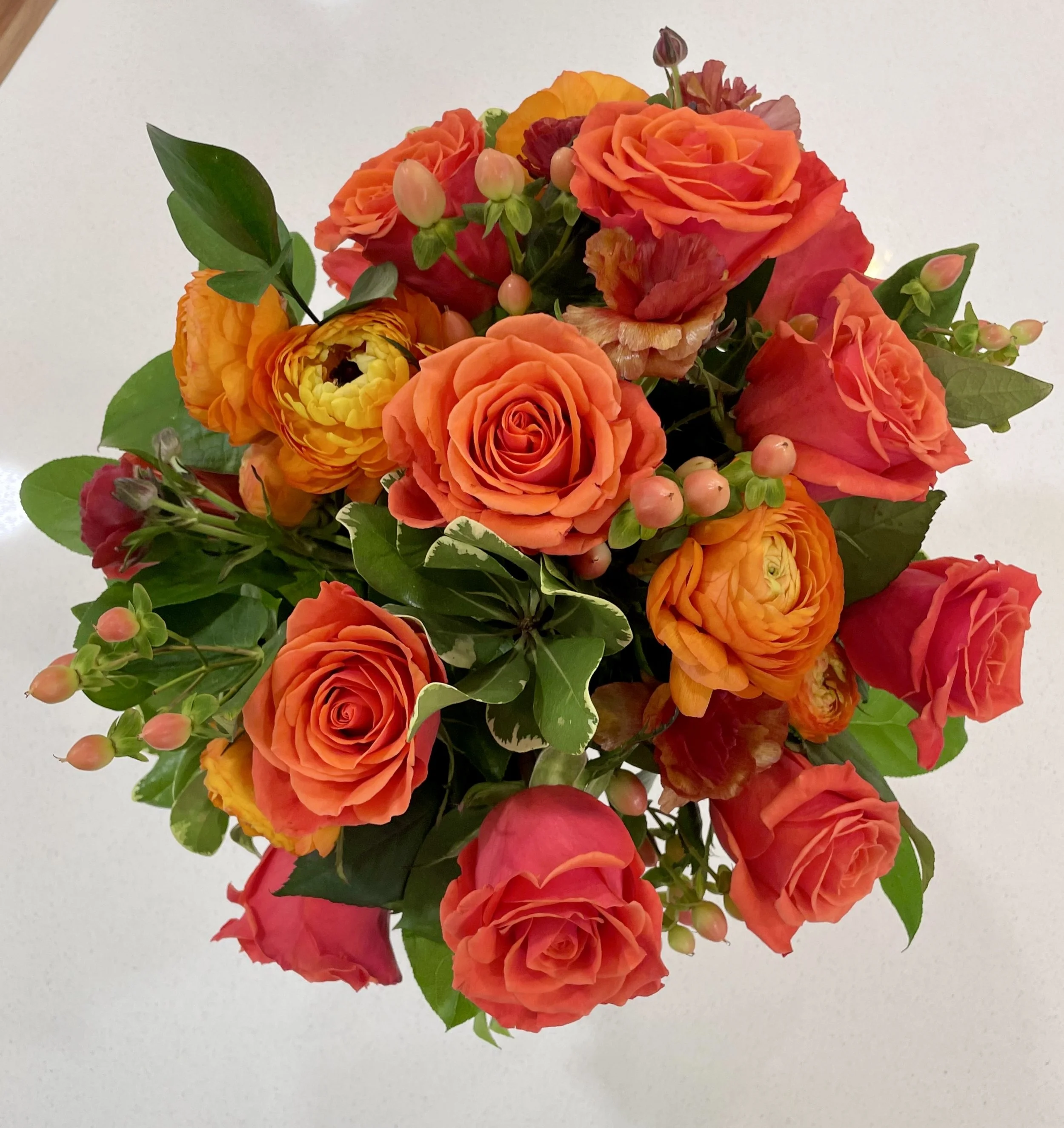 A bouquet of orange and pink roses, ranunculus, and pink berry-like accents with green leaves on a plain background.