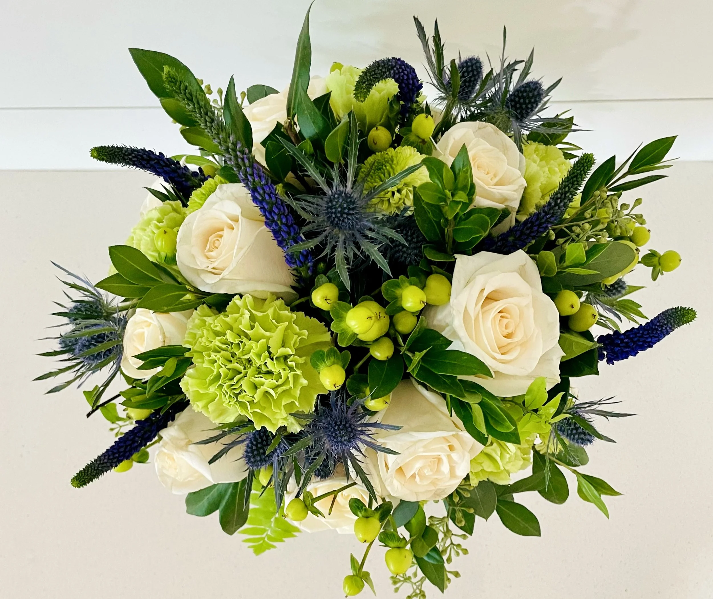 A bouquet of white roses, green carnations, blue thistle, berries, and assorted greenery.