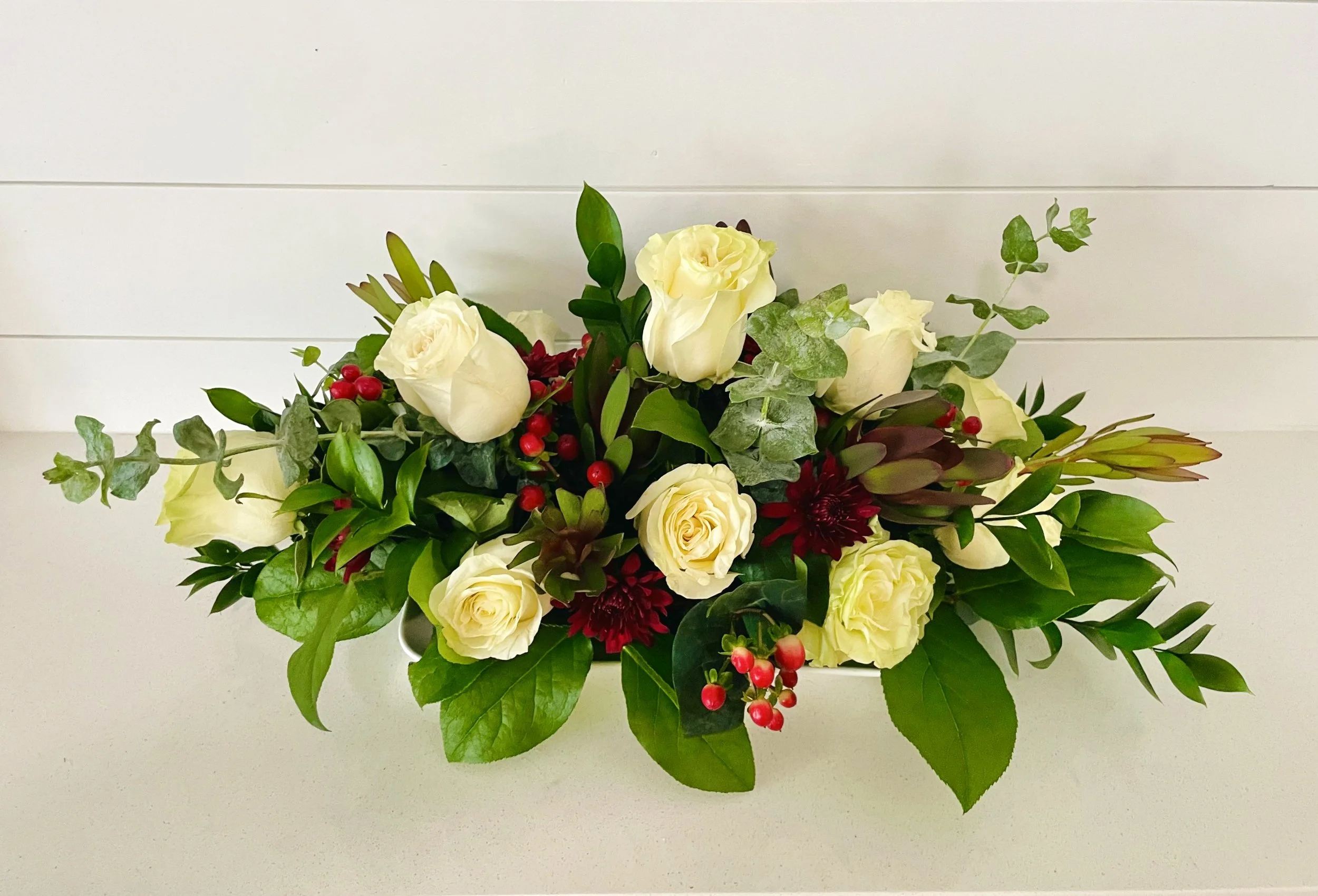 Arrangement of white roses, red berries, red flowers, and greenery on a white surface against a white paneled wall.