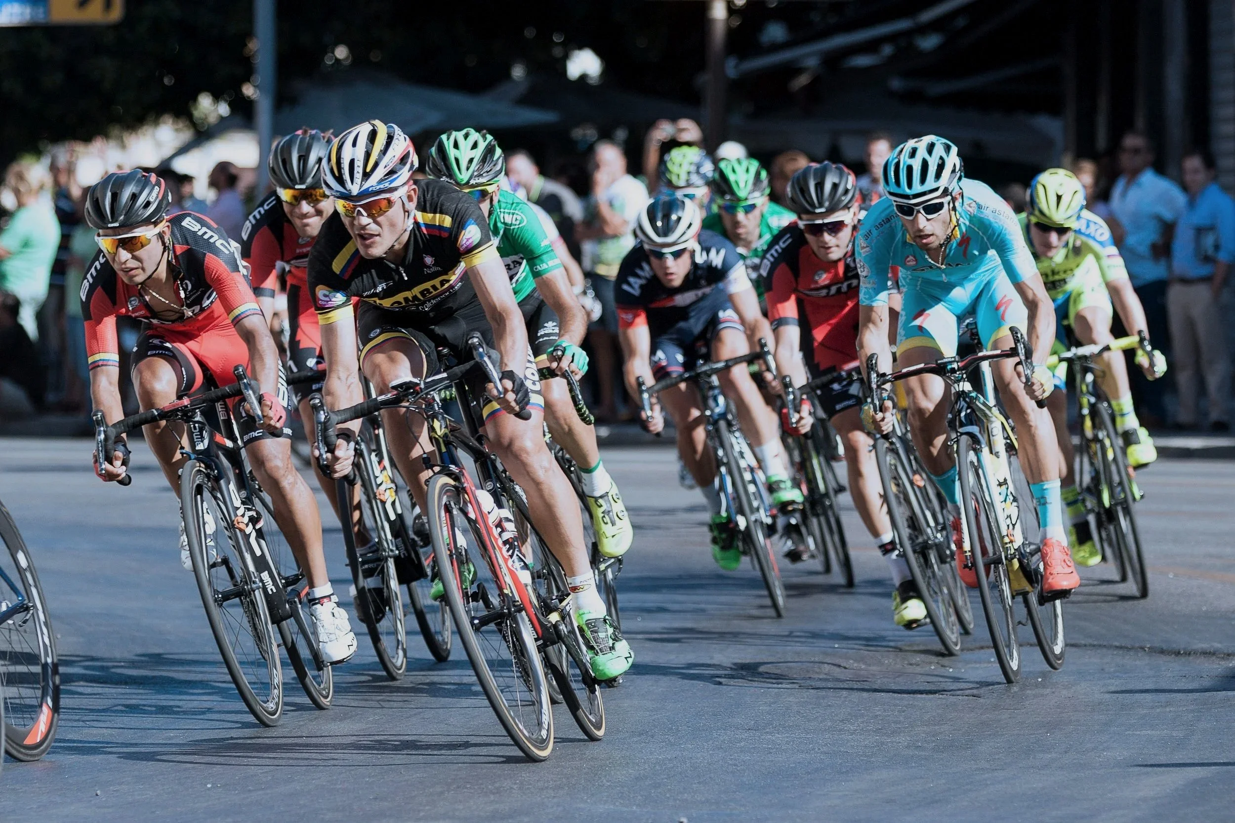 Cyclists participating in a race, wearing helmets and colorful cycling uniforms, riding closely together on a city street with spectators watching in the background.