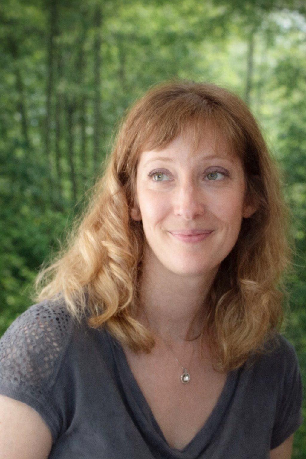 Une femme avec des cheveux ondulés clair, souriante, qui se trouve devant un fond de forêt verte.