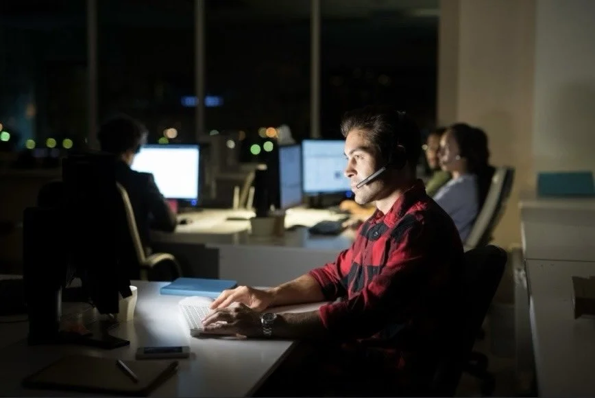 A man working at a computer in a dimly lit office during nighttime, with multiple monitors and colleagues working in the background.