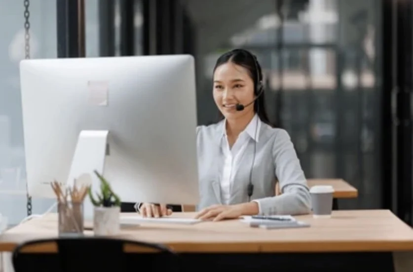 Woman working at a desk in an office, wearing a headset and smiling at the computer screen.