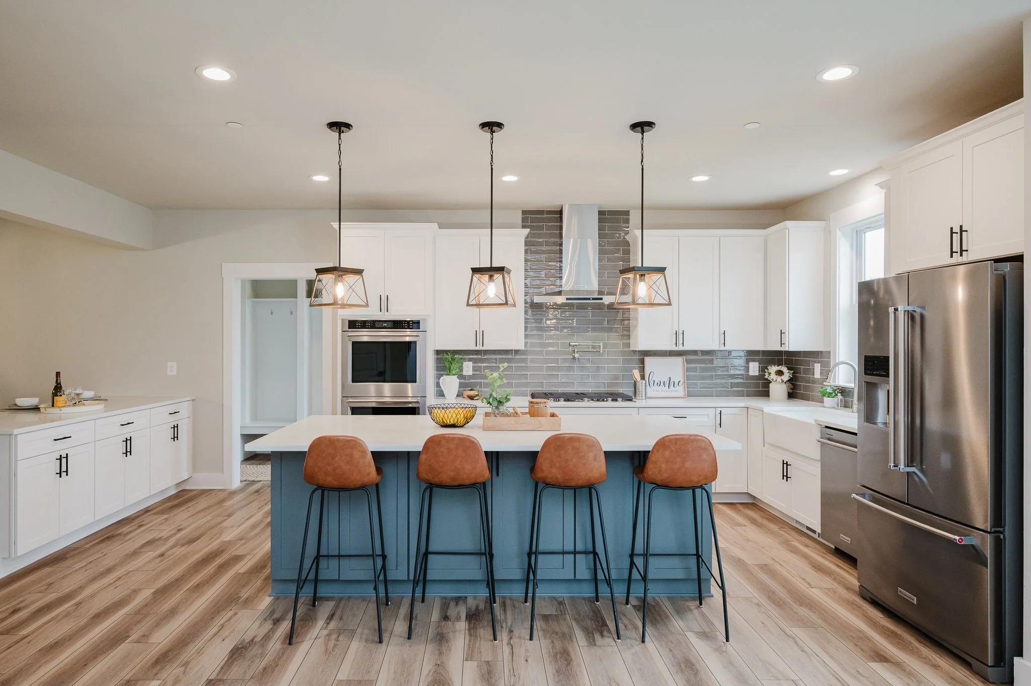 Modern kitchen with white cabinets, gray tile backsplash, stainless steel appliances, a blue kitchen island with four brown chairs, hardwood floors, and pendant lighting.