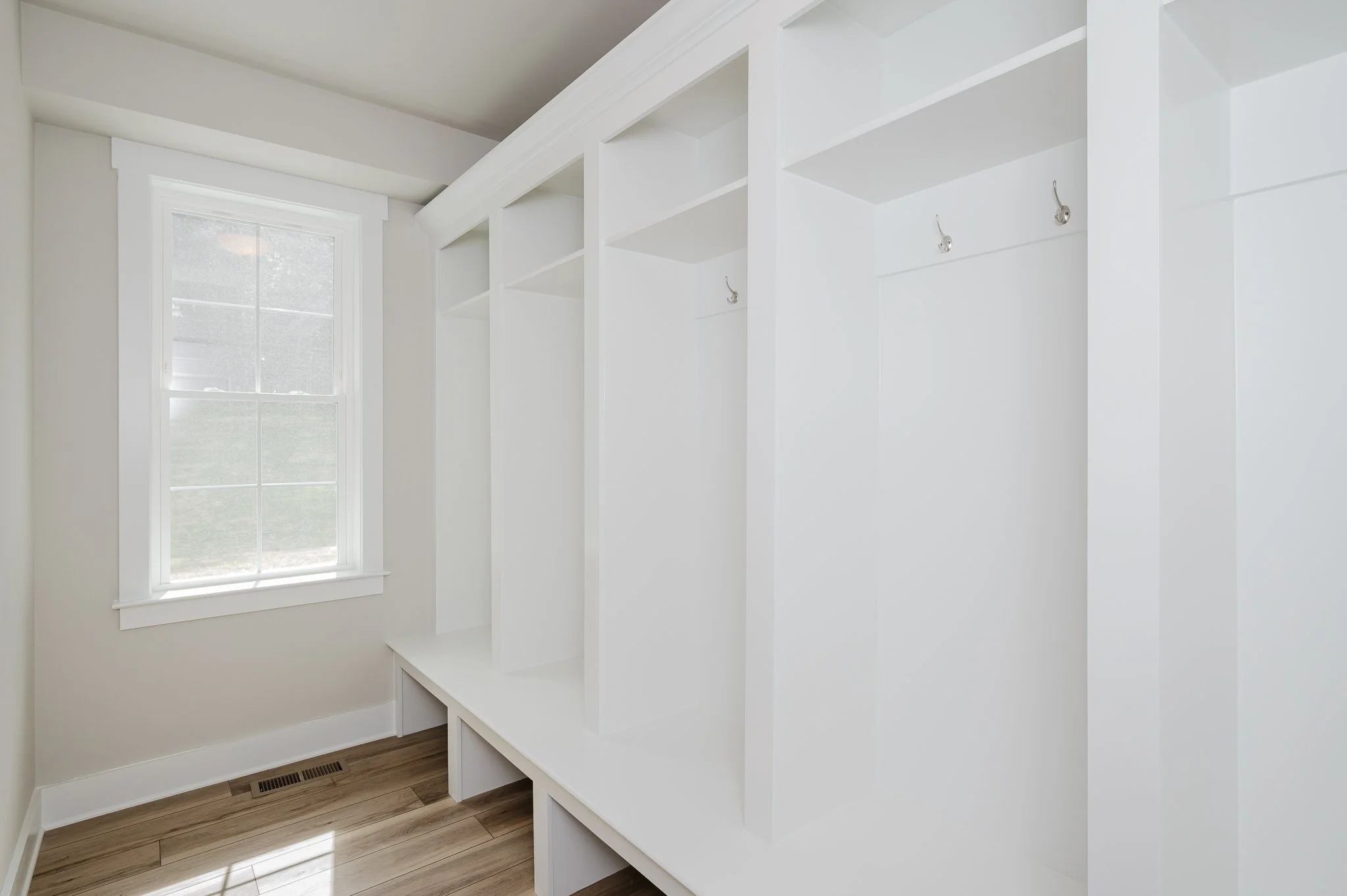 Empty white closet with hooks, built-in shelving, and a window in a room with hardwood floors.