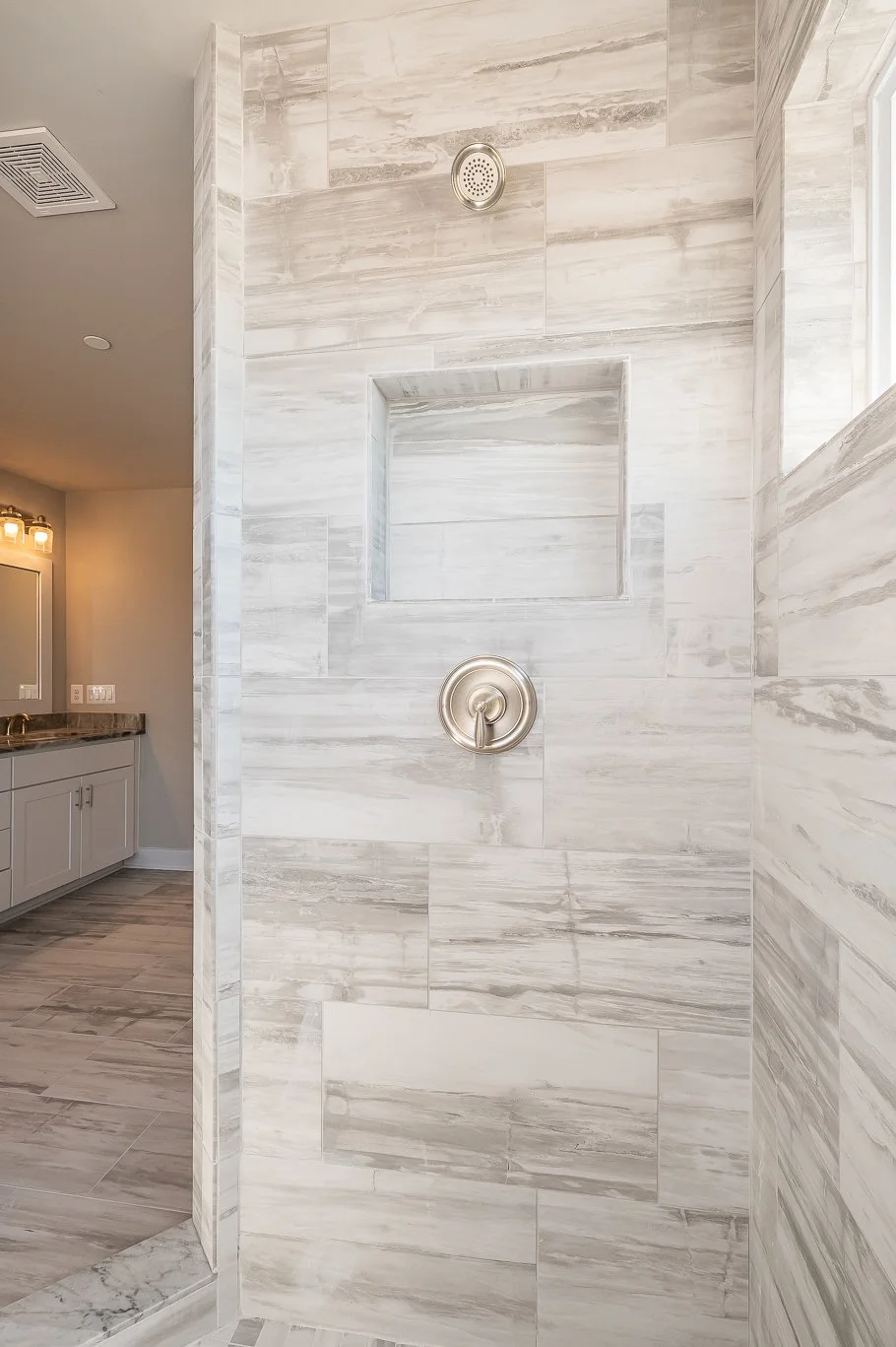 A modern shower with light gray marble tiles, a built-in niche, a chrome showerhead, and a round control valve. Part of a bathroom with a vanity and mirror is visible in the background.