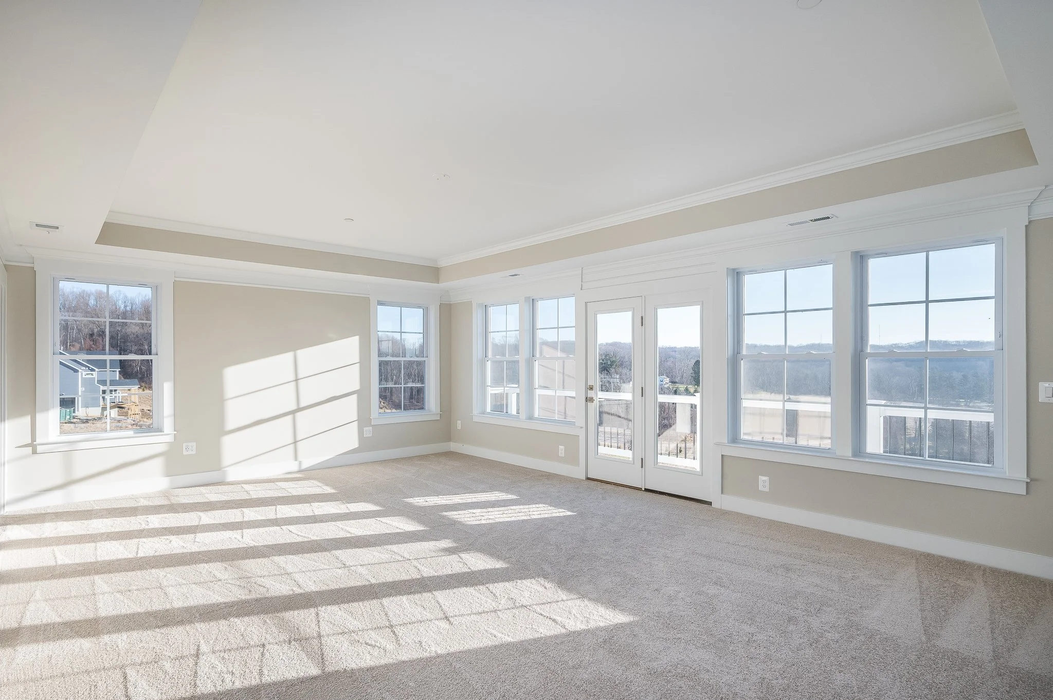 Empty room with beige carpet, multiple large windows, and a glass door leading outside, with sunlight casting shadows.