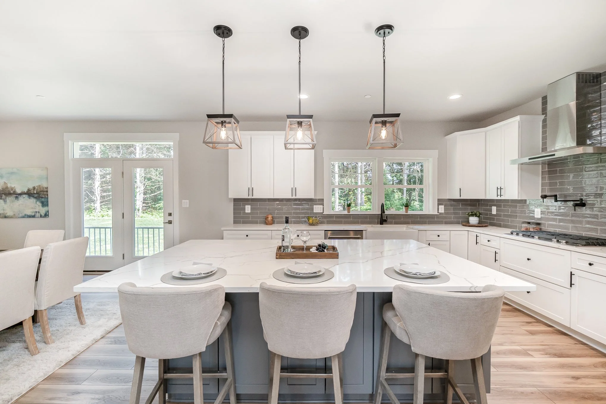 Modern kitchen with white cabinets, gray tile backsplash, and a large white marble island with four beige upholstered bar stools. Three pendant lights hang above the island, and there are windows letting in natural light.