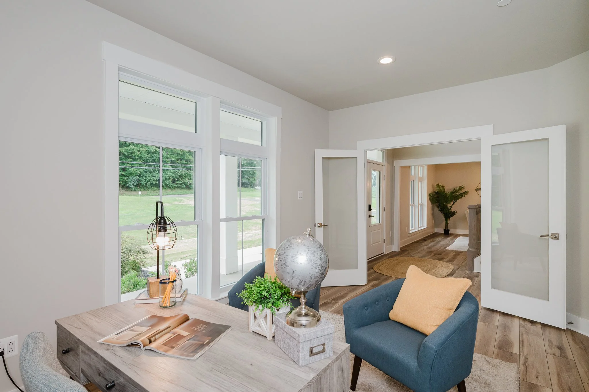 Living room with large windows, a desk with a globe, a blue armchair with a yellow pillow, and an open doorway leading to another room with a potted plant.