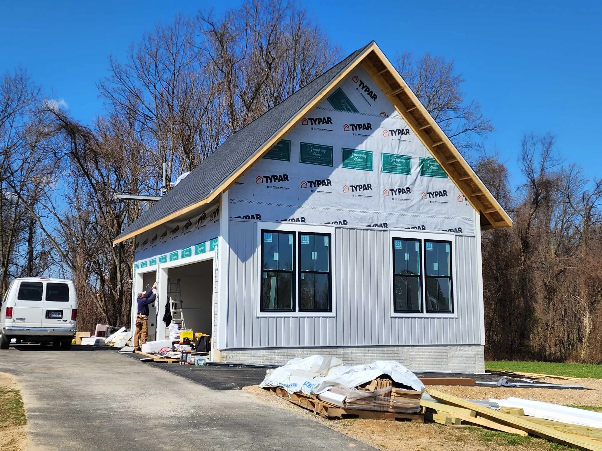 A beige garage building with two garage doors, a small window above, and a central door with a flower pot in front. There is a paved driveway and a backyard with a pool, a fence, and greenery under a partly cloudy sky.