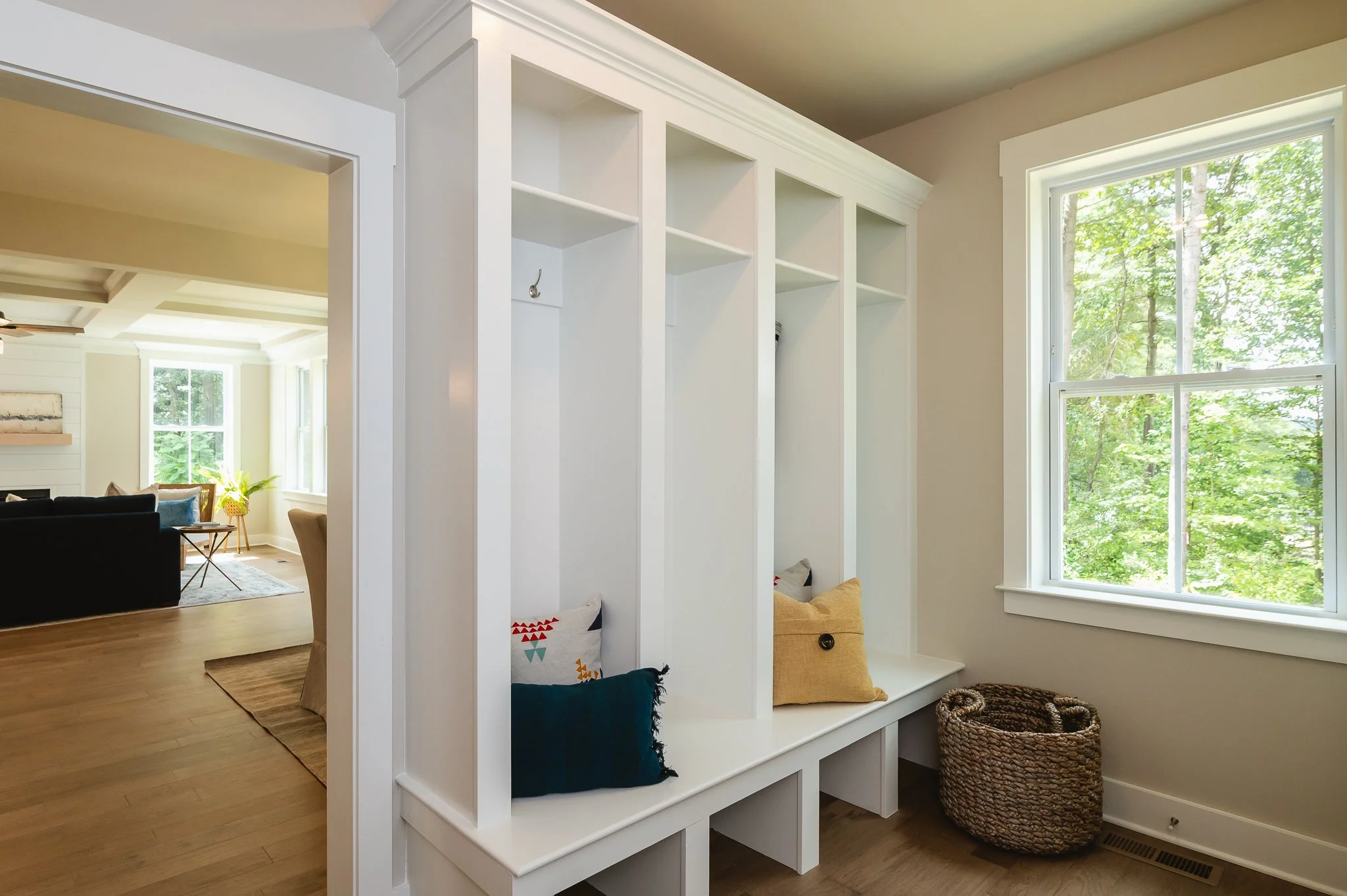 Hallway with built-in white cubbies with cushions and a woven basket, next to a window with green foliage outside.