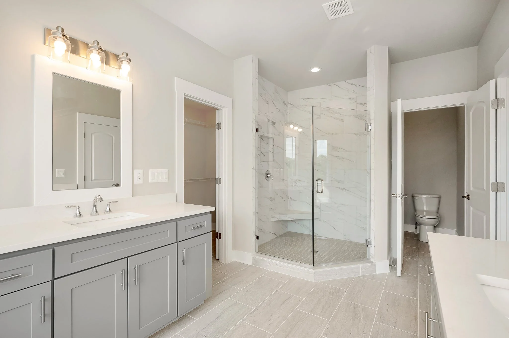 Modern bathroom with light gray vanity, large mirror, shower with glass doors, small toilet area, and beige tiled floors and walls.