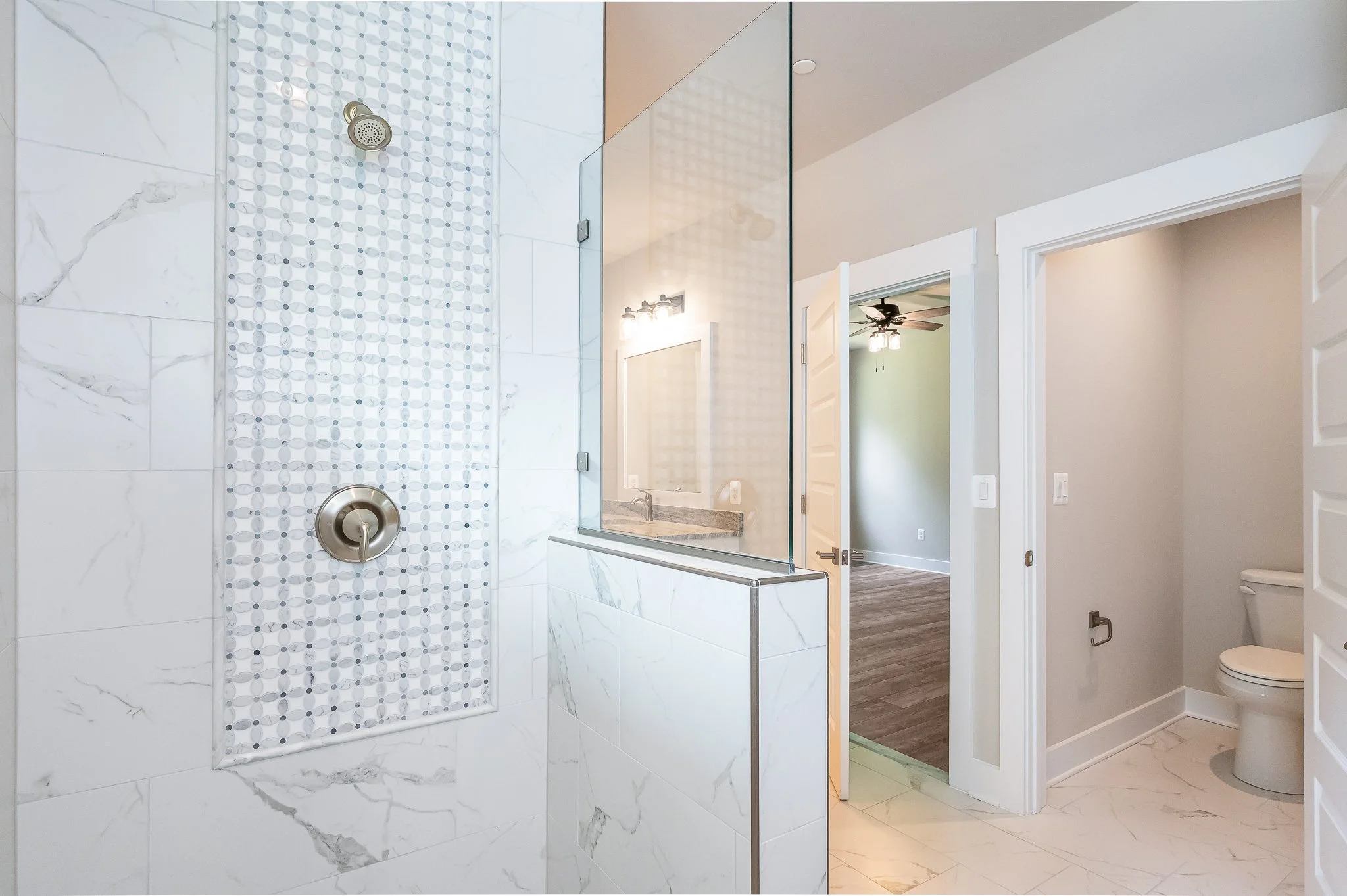Modern bathroom with white marble tiles, walk-in shower with glass door, and adjacent toilet area.