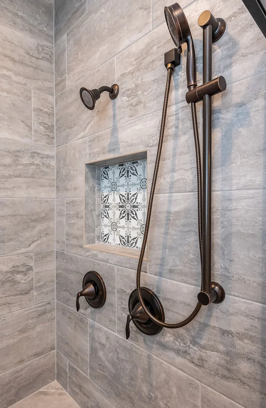A modern shower with a handheld showerhead and a fixed showerhead, both in bronze finish, installed on a gray tiled wall with a decorative tile inside a shower niche.