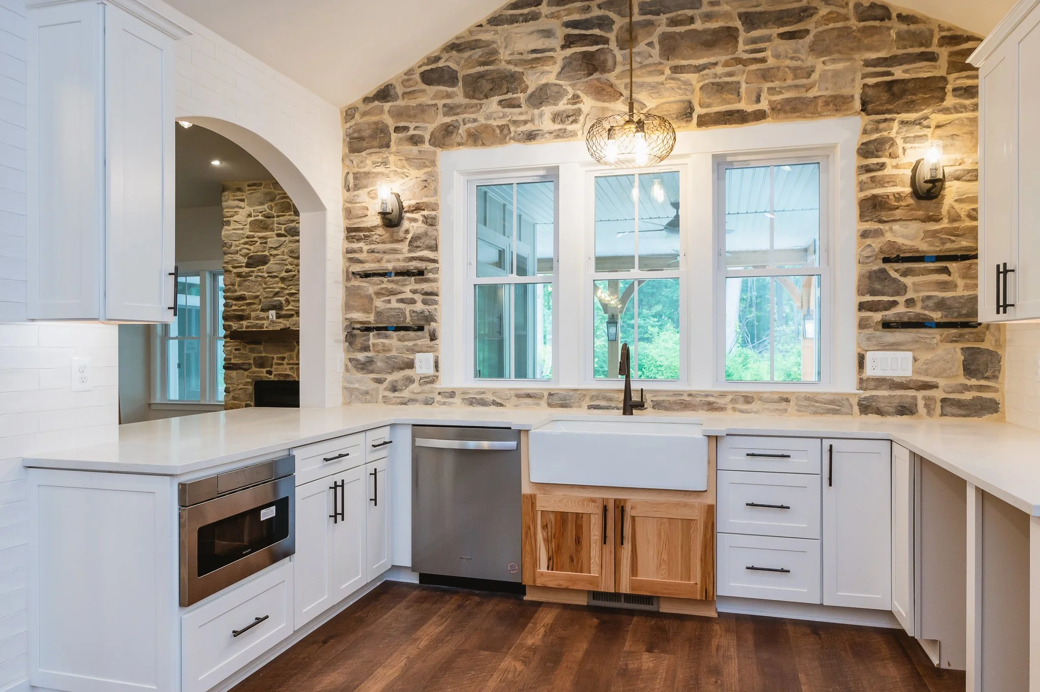 Kitchen with white cabinets, wooden flooring, stone accent wall, large window above sink, and modern lighting fixtures.