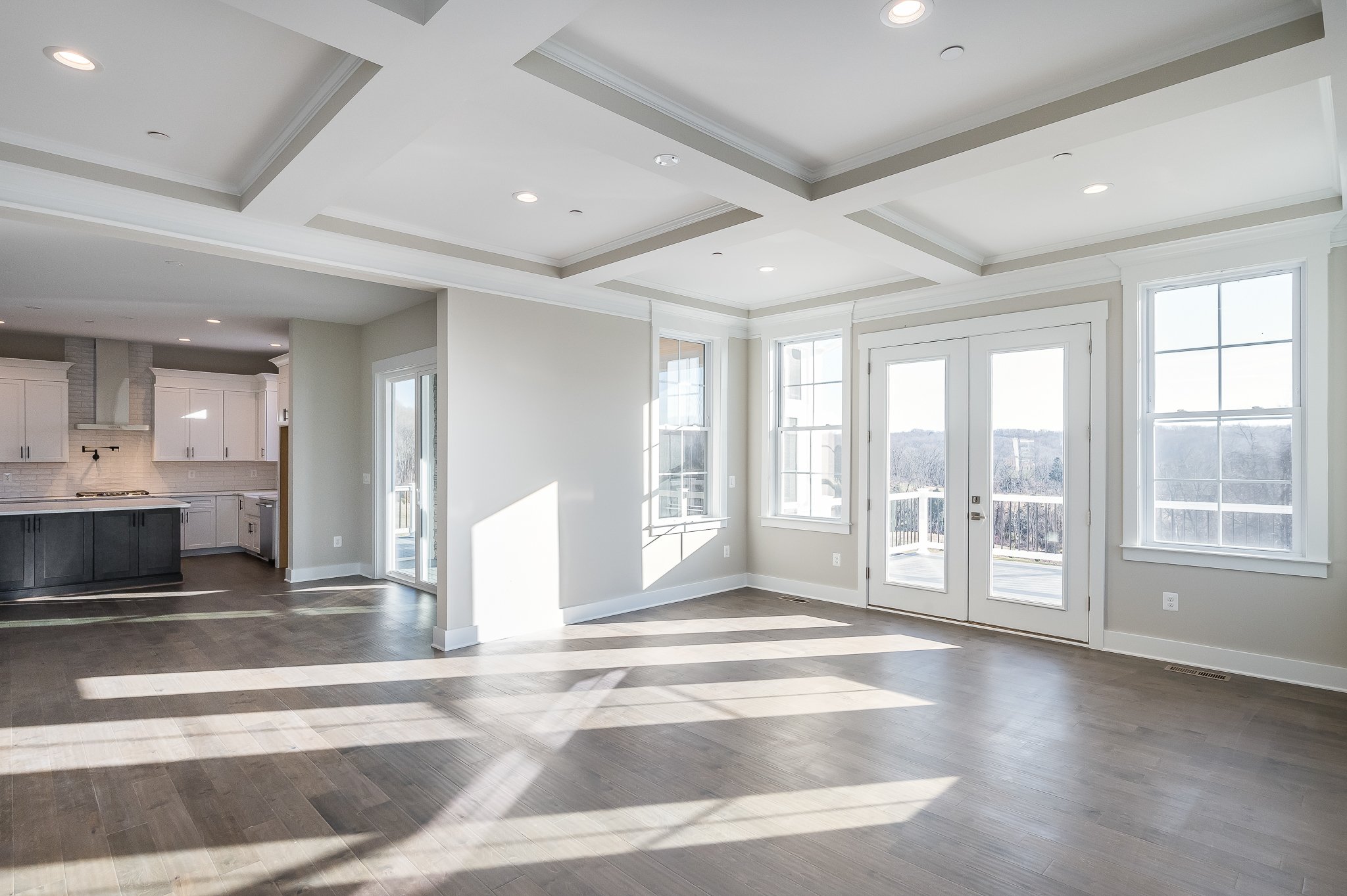 Bright empty living room with large windows and glass doors, hardwood floors, and tray ceiling, adjacent to a modern kitchen with white cabinets and dark island.