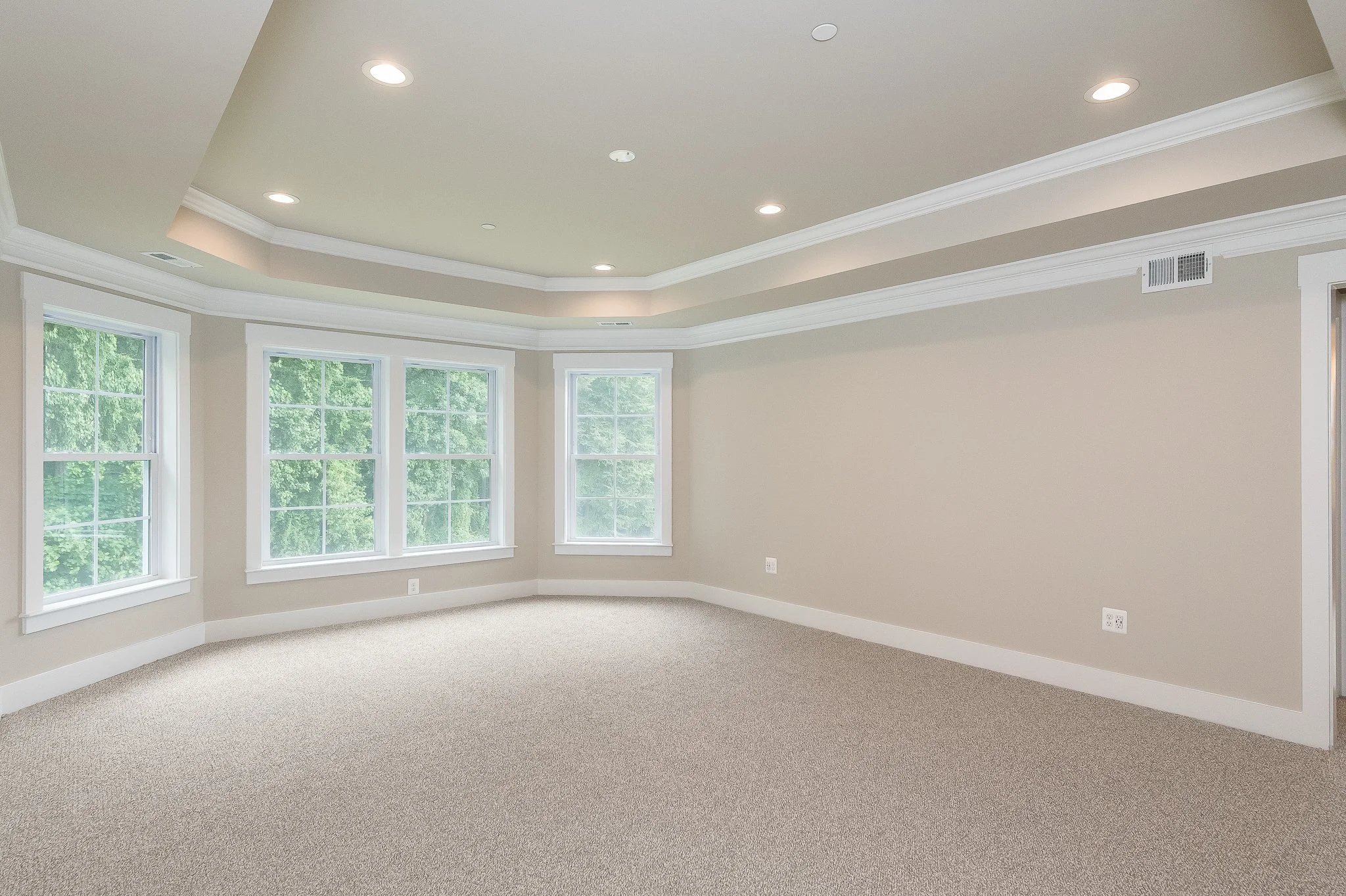 Empty living room with large windows, beige walls, white trim, and beige carpet flooring.
