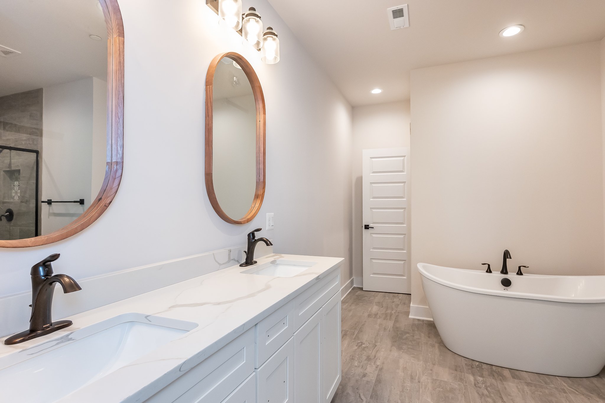 Modern bathroom with a double vanity with white marble countertop, two oval mirrors with wooden frames, and a standalone white bathtub. The bathroom has beige walls, gray tiled floor, and recessed lighting.