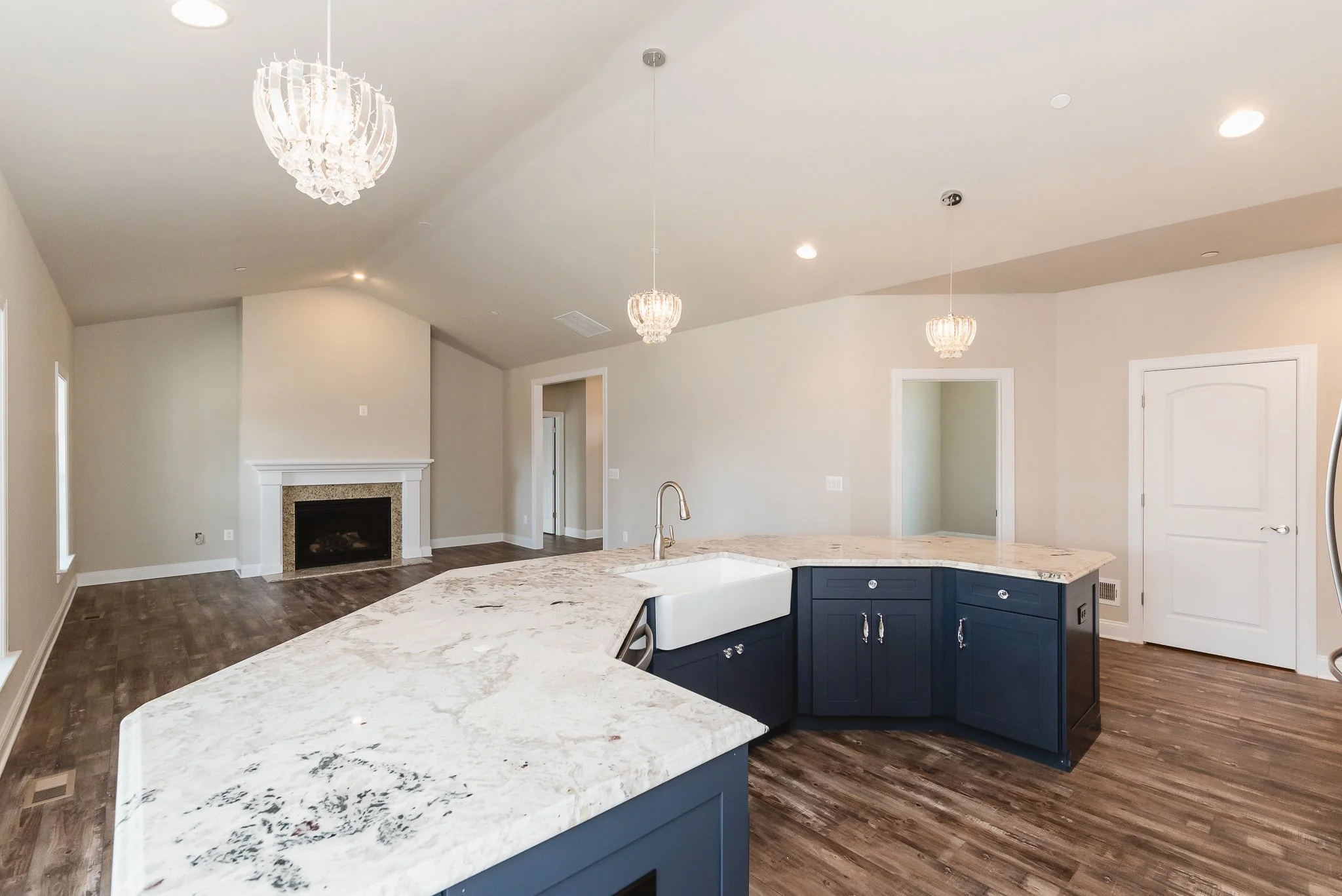 Open-concept kitchen and living room with hardwood floors, blue kitchen cabinets, a large marble island, white farmhouse sink, chandelier lighting, fireplace, and doorways leading to other rooms.