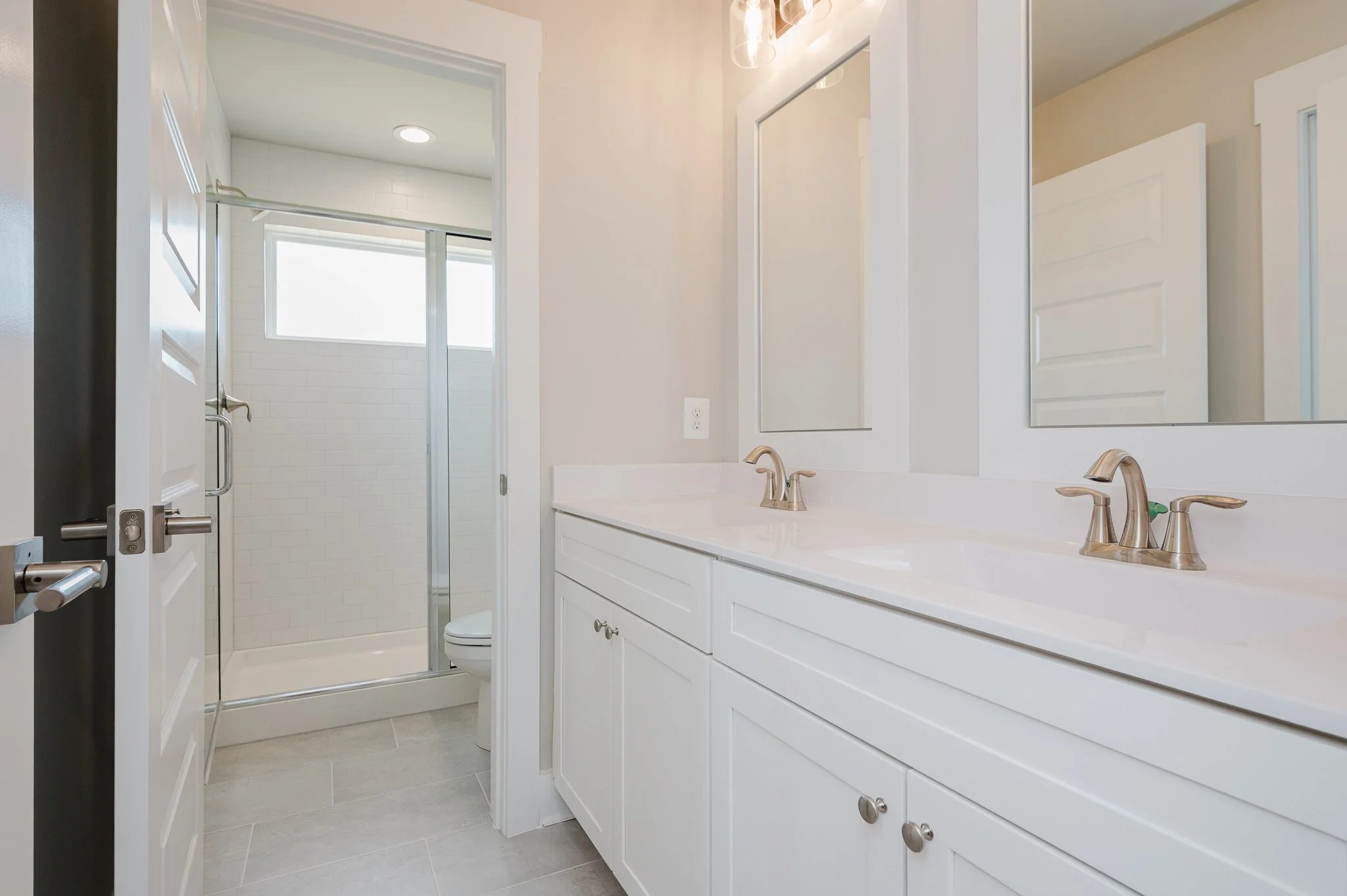 White bathroom vanity with dual sinks and mirrors, white cabinets, and a shower stall with a sliding glass door and white tiled walls, with a small window above.