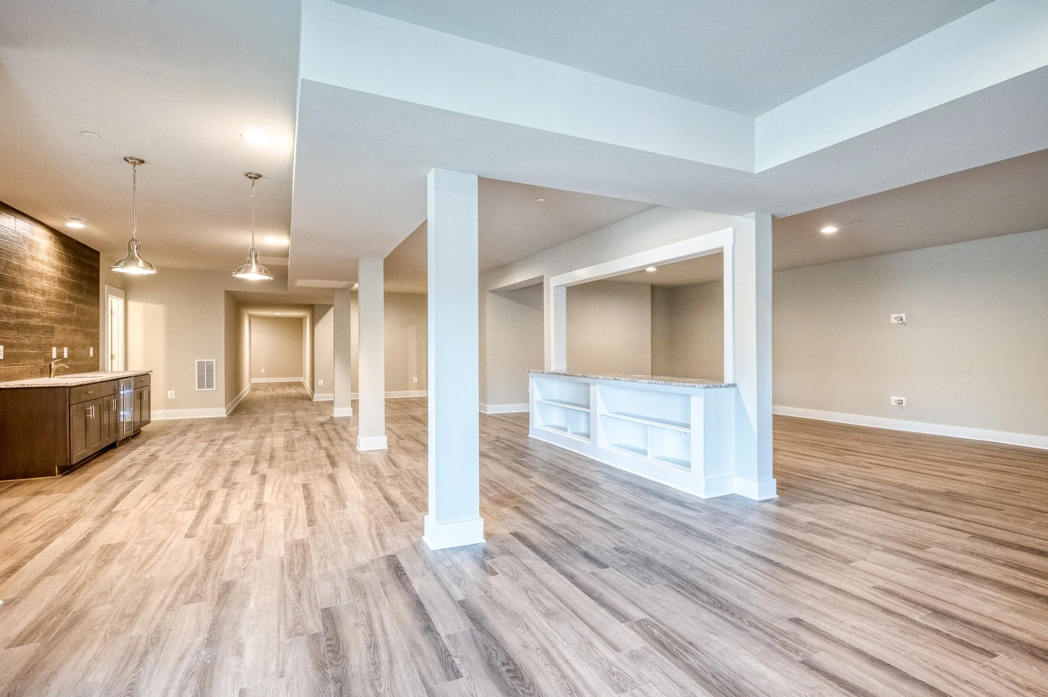 Empty open-concept living space with wood flooring, white walls, a built-in white shelf, and a kitchen area with dark wood cabinets and pendant lighting.