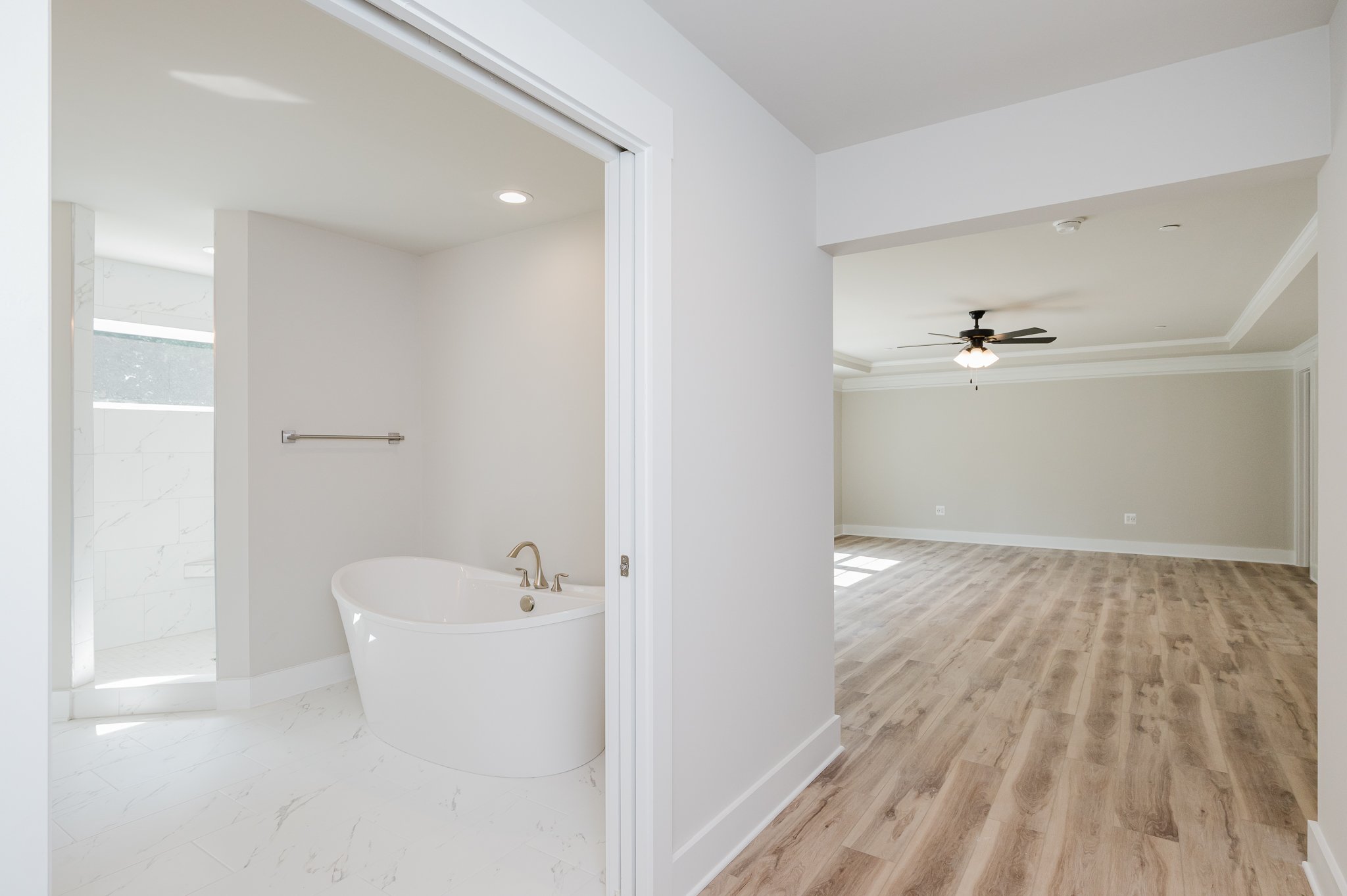 Empty modern living room with light wood flooring, ceiling fan, and neutral-colored walls. Adjacent to it, a bathroom with a white bathtub and marble flooring is visible through an open sliding door.
