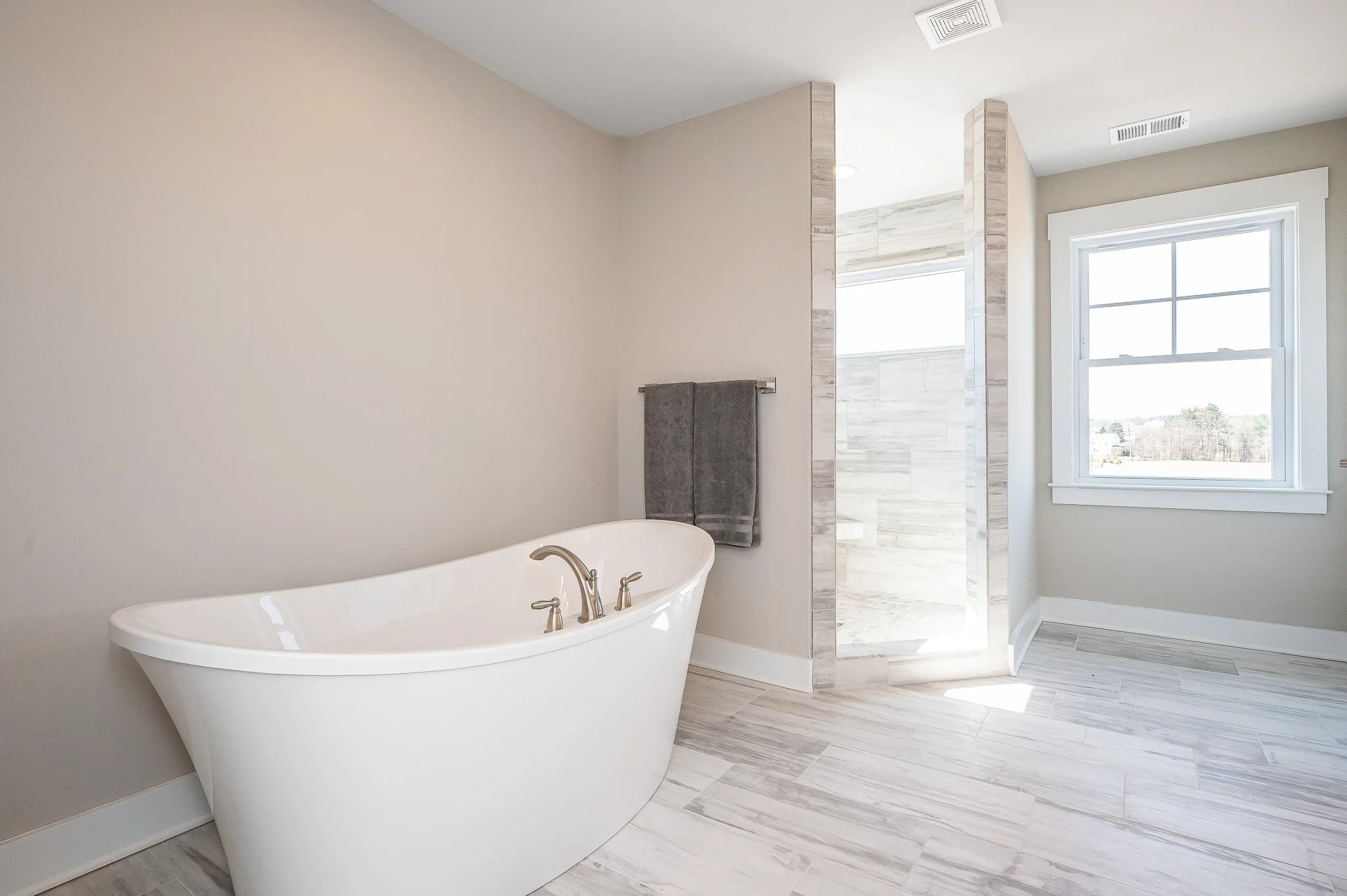 A modern bathroom featuring a white freestanding bathtub, a gray towel hanging on a rack, a large window, and a shower area with light-colored tiled walls.