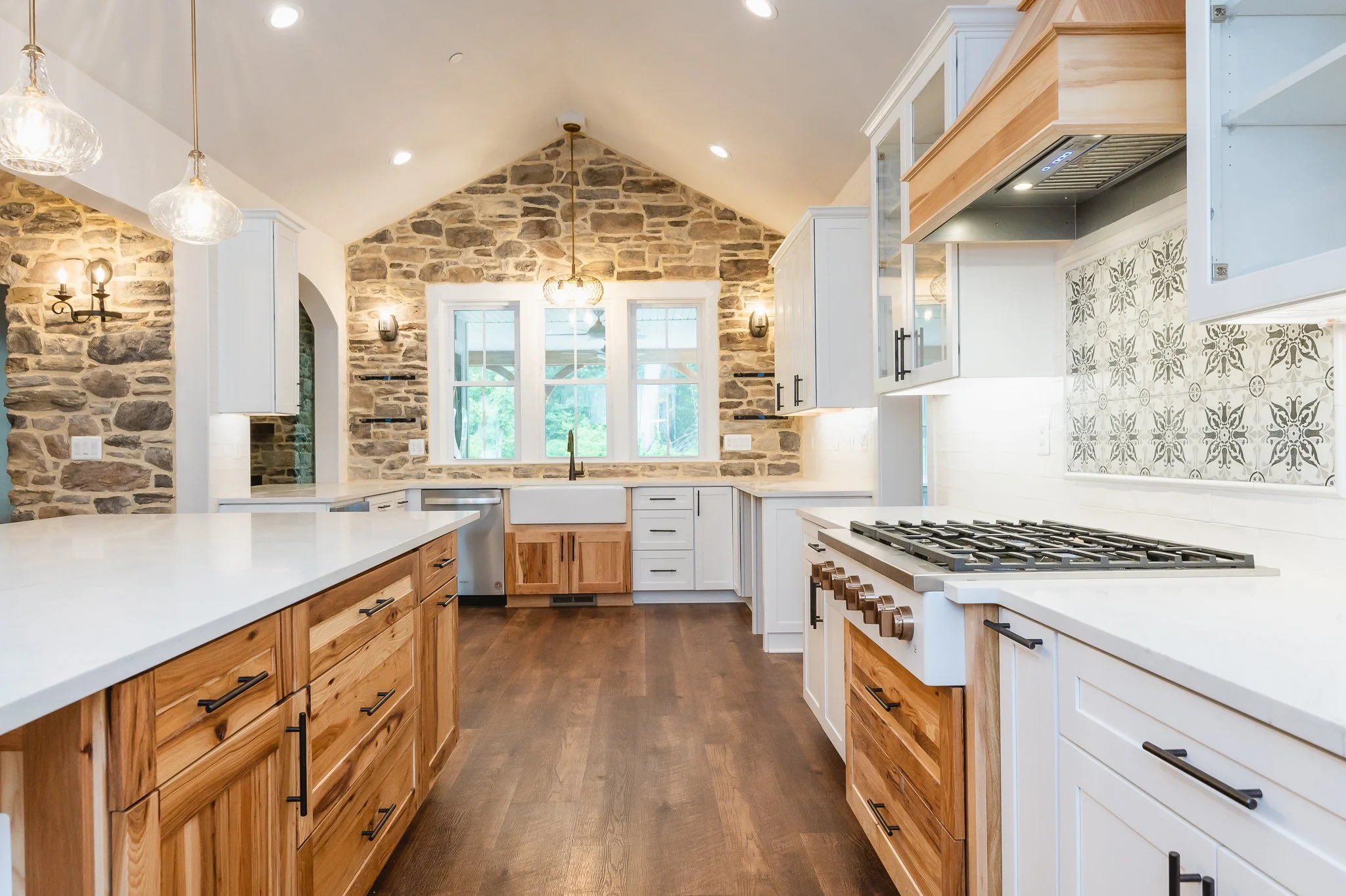 Kitchen with white cabinets, wooden drawers, a white countertop, a stone accent wall, a window above the sink, patterned tiles behind the stove, and overhead lighting.