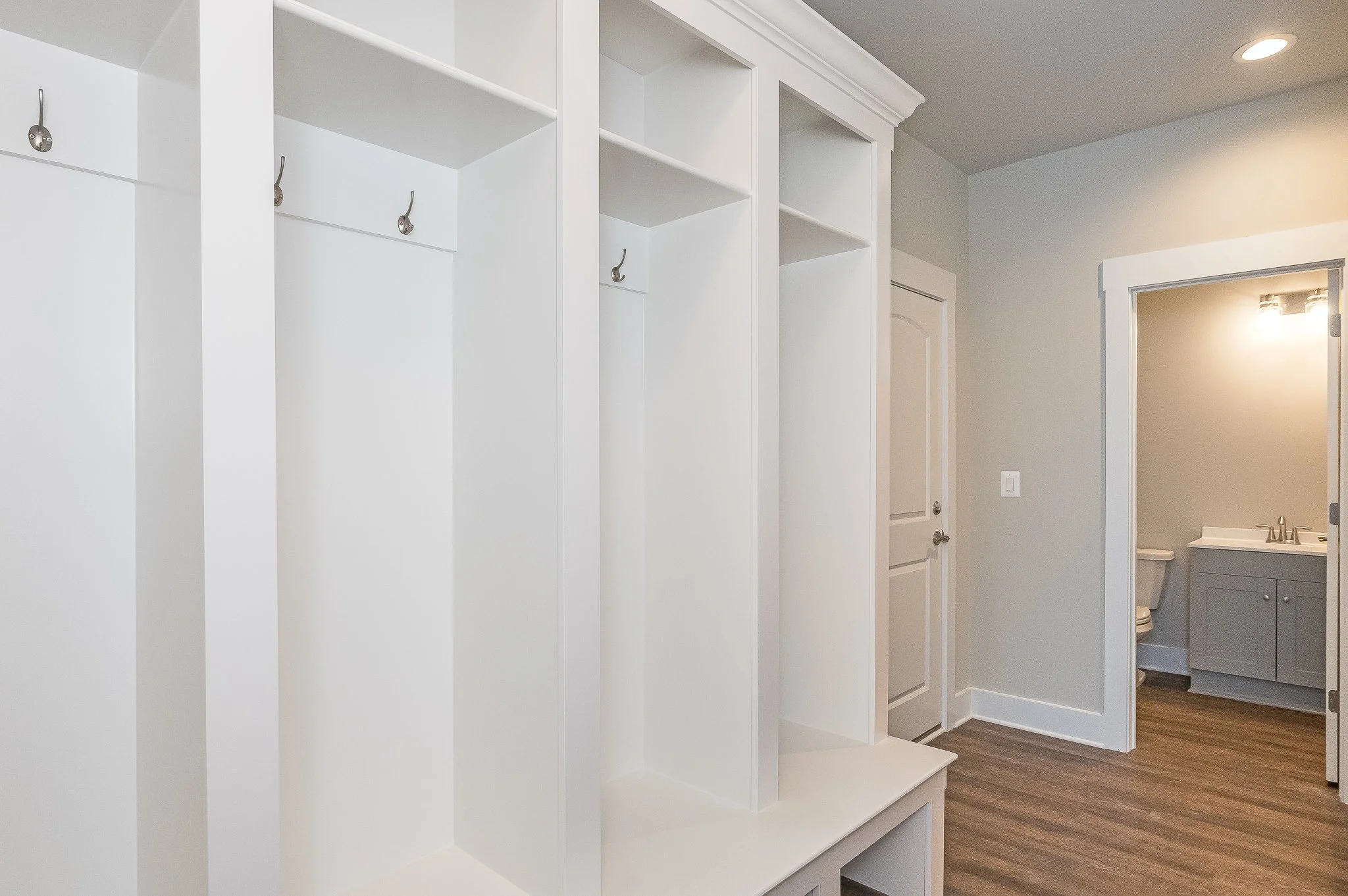 Empty white built-in closet with hooks and shelves, adjacent to a doorway leading to a bathroom with a vanity, toilet, and gray walls, in a modern home interior.