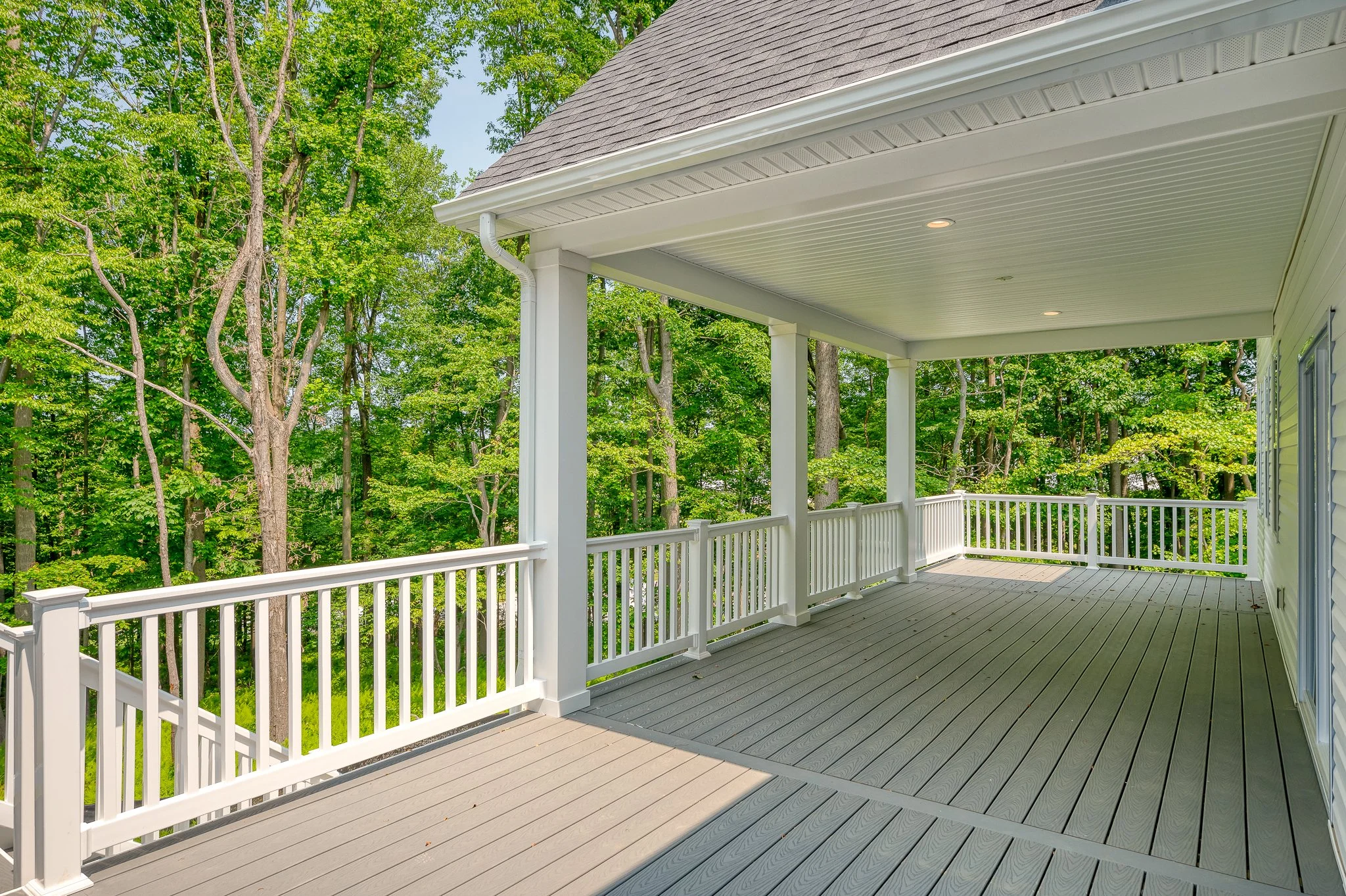 View of a spacious outdoor balcony with white railings and columns, overlooking lush green trees on a bright, sunny day.