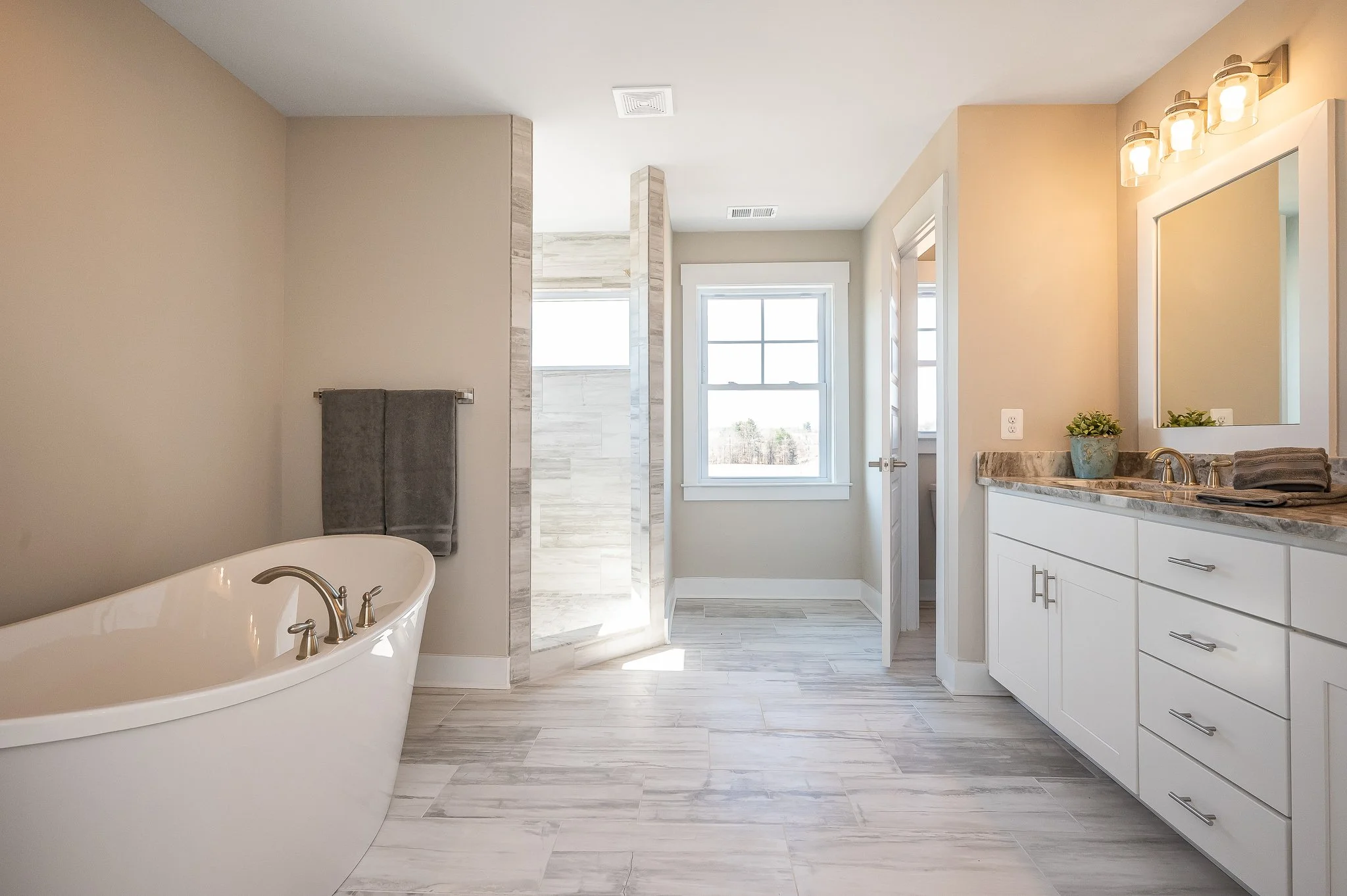 Bright bathroom with a white bathtub, a glass shower enclosure, a window, and a white vanity with a marble countertop and a mirror