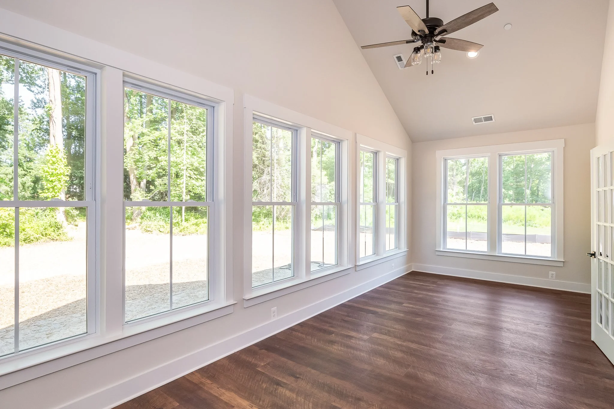 Empty room with large windows overlooking a green outdoor area, with dark wooden flooring and a ceiling fan.