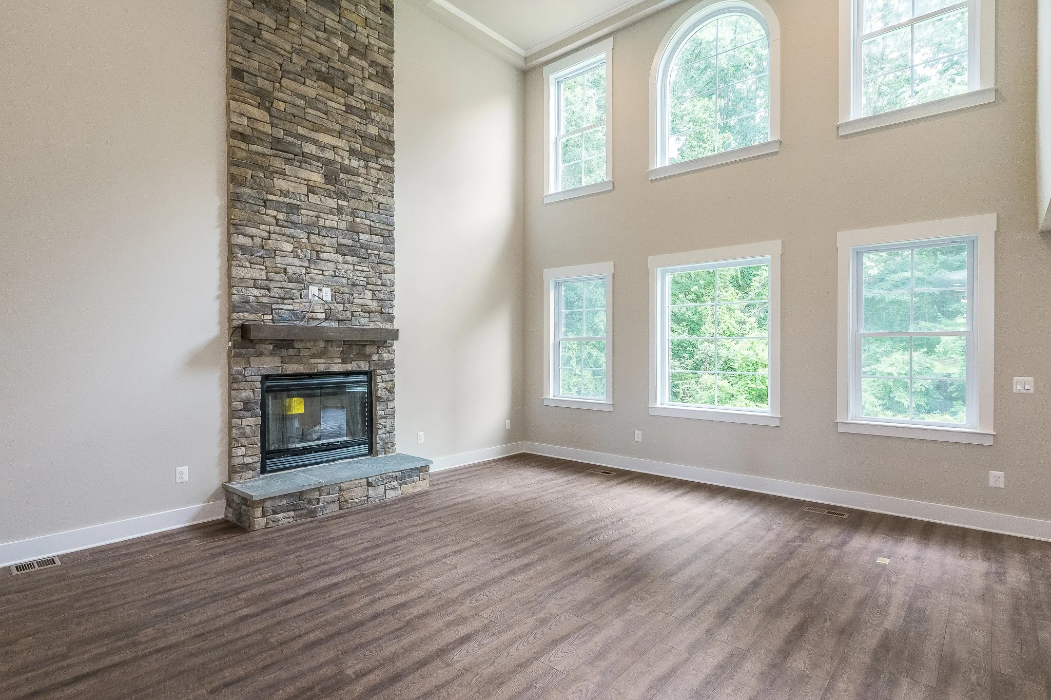 Empty living room with a stone fireplace, four large windows, and hardwood flooring.