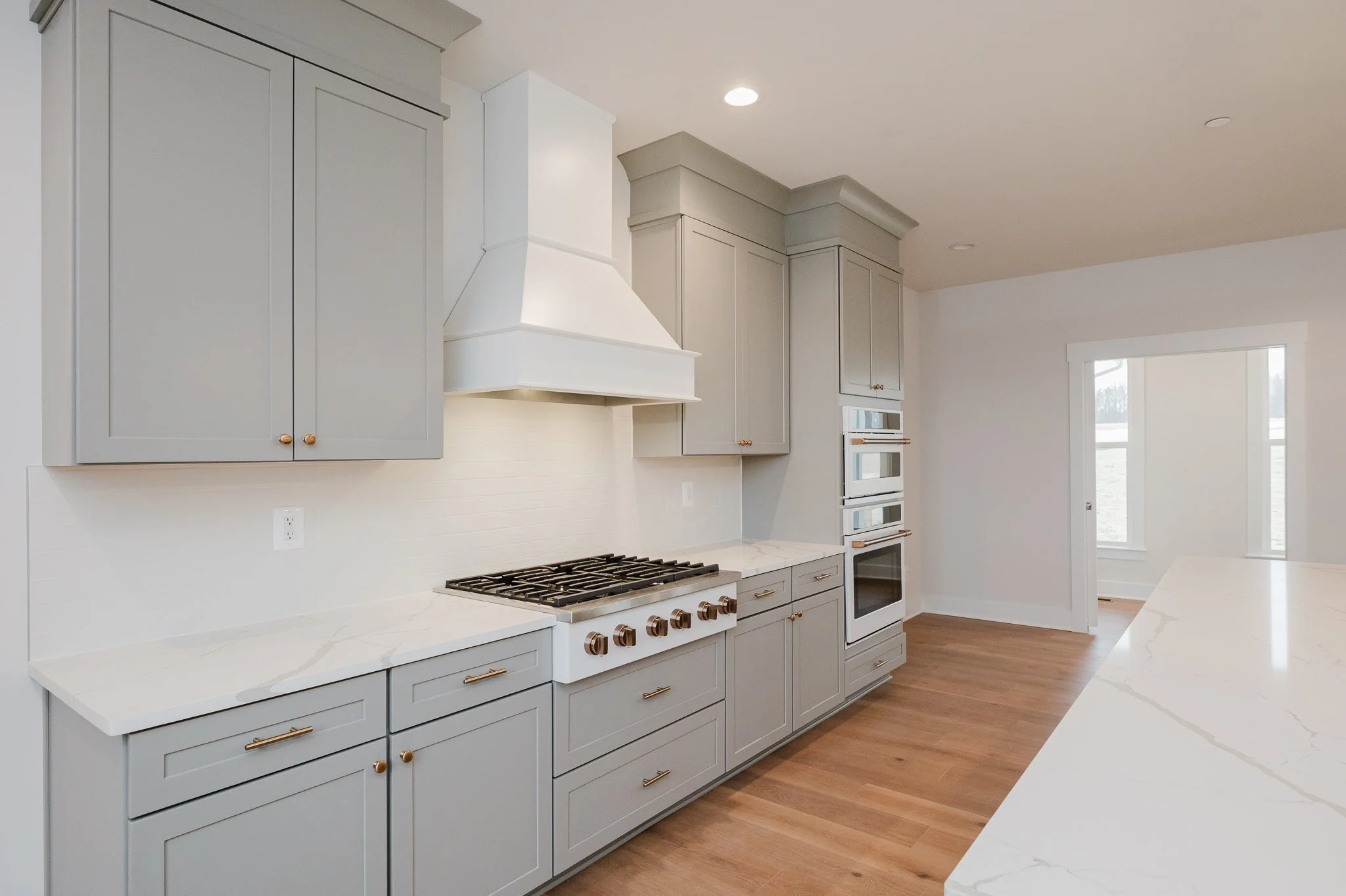 A modern kitchen with light gray cabinets, white marble countertops, and a wooden floor. There is a cooktop, wall-mounted oven, and microwave, with a white range hood above the stove. Natural light enters through a window and a door in the background