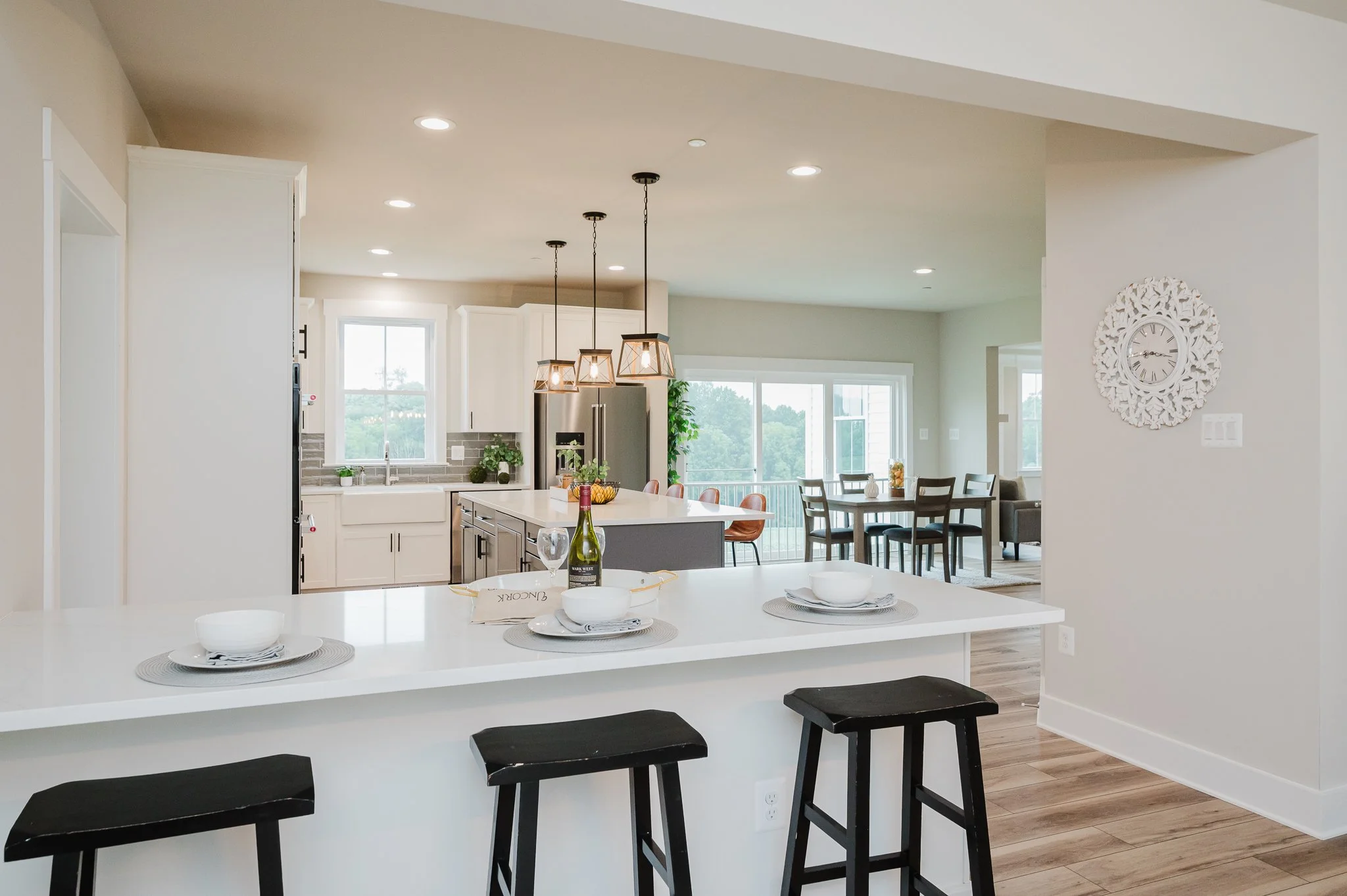 Open-concept kitchen and dining area with white cabinetry, stainless steel refrigerator, window above the sink, pendant lights over the island, and a dining table with chairs in the background.