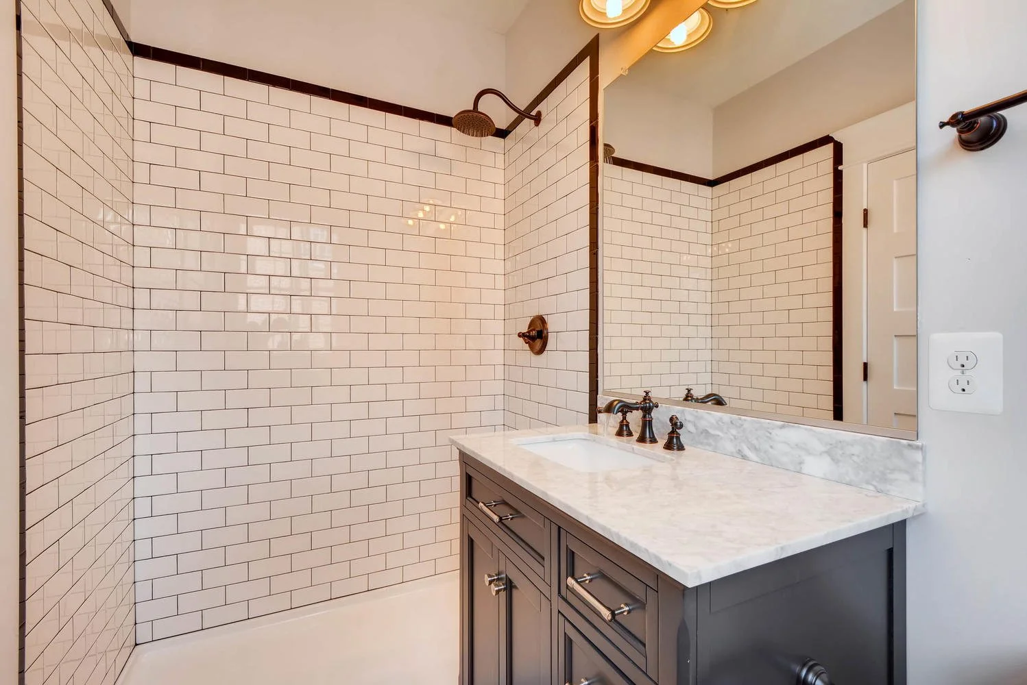 Bathroom with white subway tile walls, a shower area with a rainfall showerhead, a dark wood vanity with a marble countertop, and a large mirror.