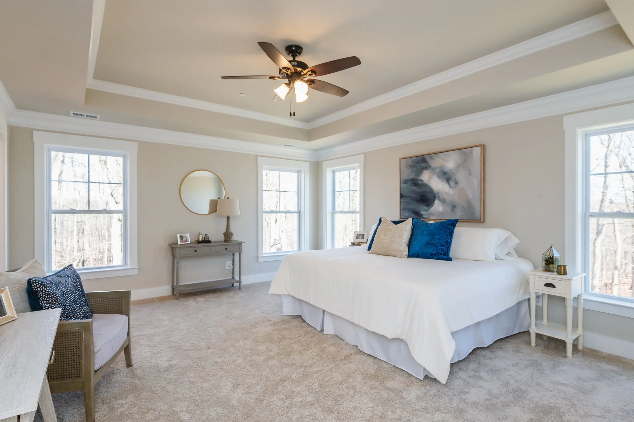 A spacious bedroom with a large bed, blue and beige pillows, white bedding, and a nightstand with a small decorative item. There are three windows with natural light, a ceiling fan, a gray console table with a mirror and lamp, and beige carpet floori