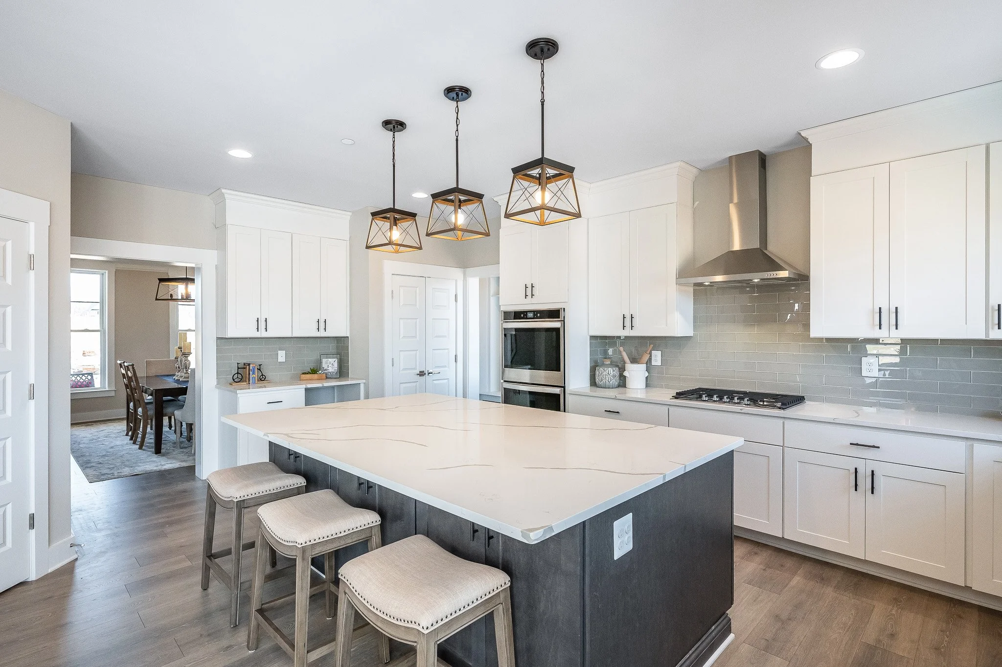 Modern kitchen with white cabinets, gray subway tile backsplash, stainless steel range hood and oven, central island with white marble countertop, three pendant lights, and three beige barstools.