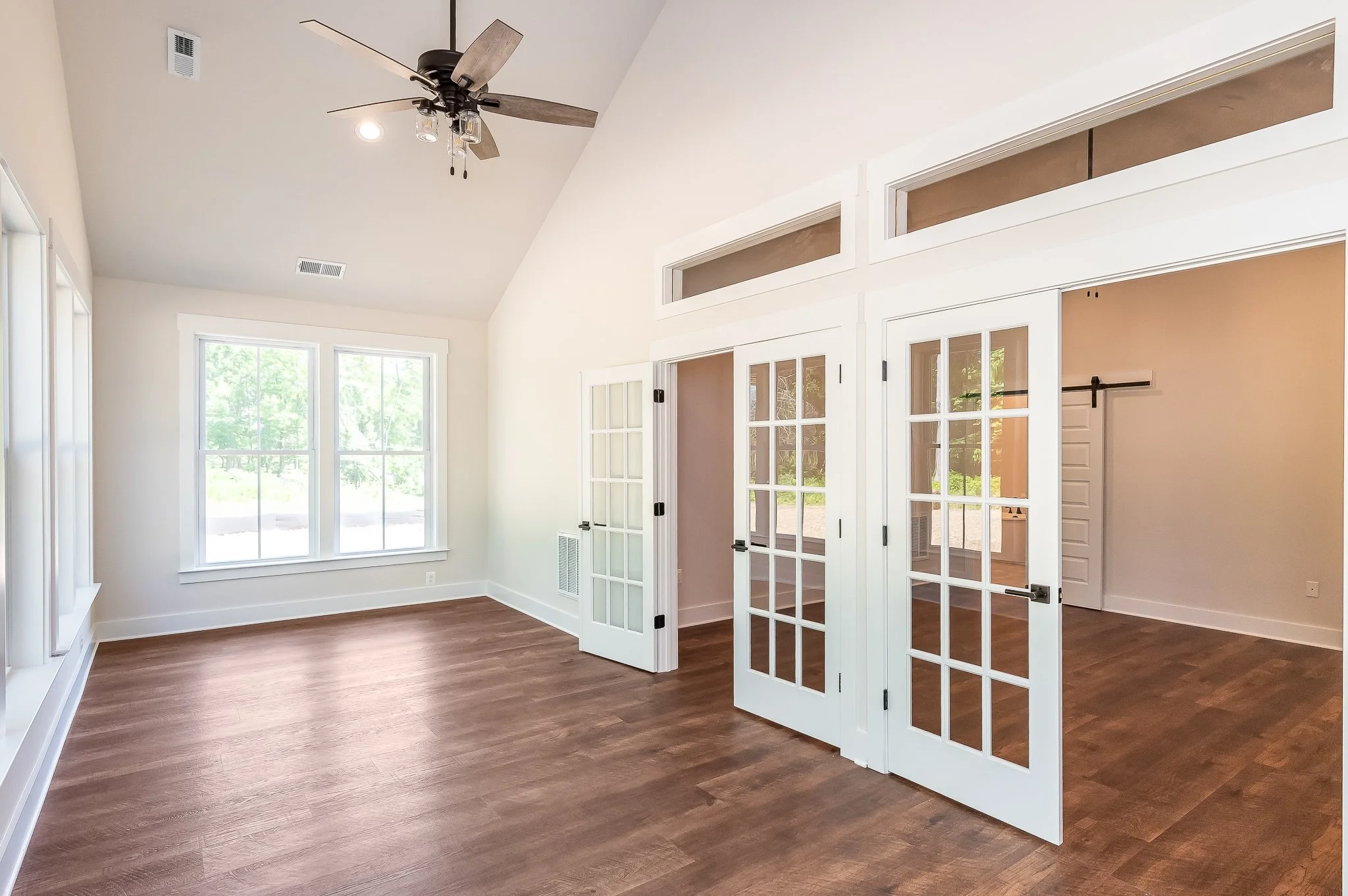 Empty room with large window, wooden floors, white walls, and french doors leading to another room.