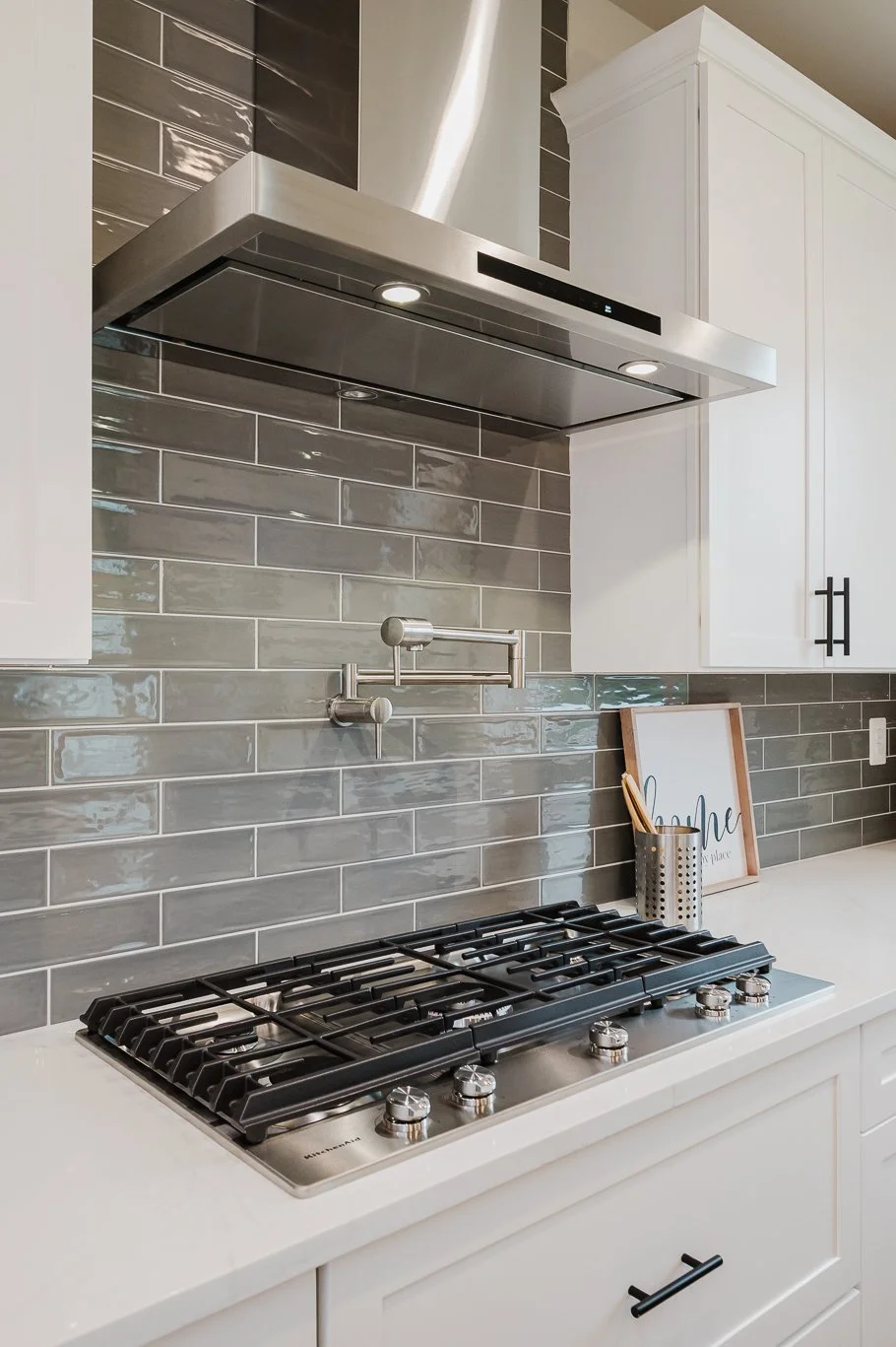 Modern kitchen with gray subway tile backsplash, white cabinets, stainless steel range hood, gas stove, and countertop with decorative items.