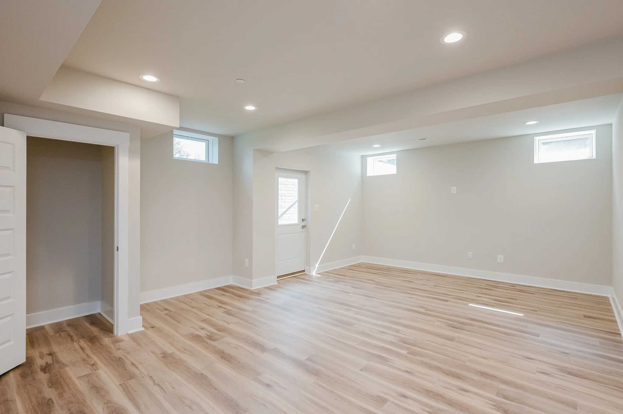 Empty room with natural light, light-colored hardwood floors, white walls, small high windows, a door with a window, and recessed ceiling lights.