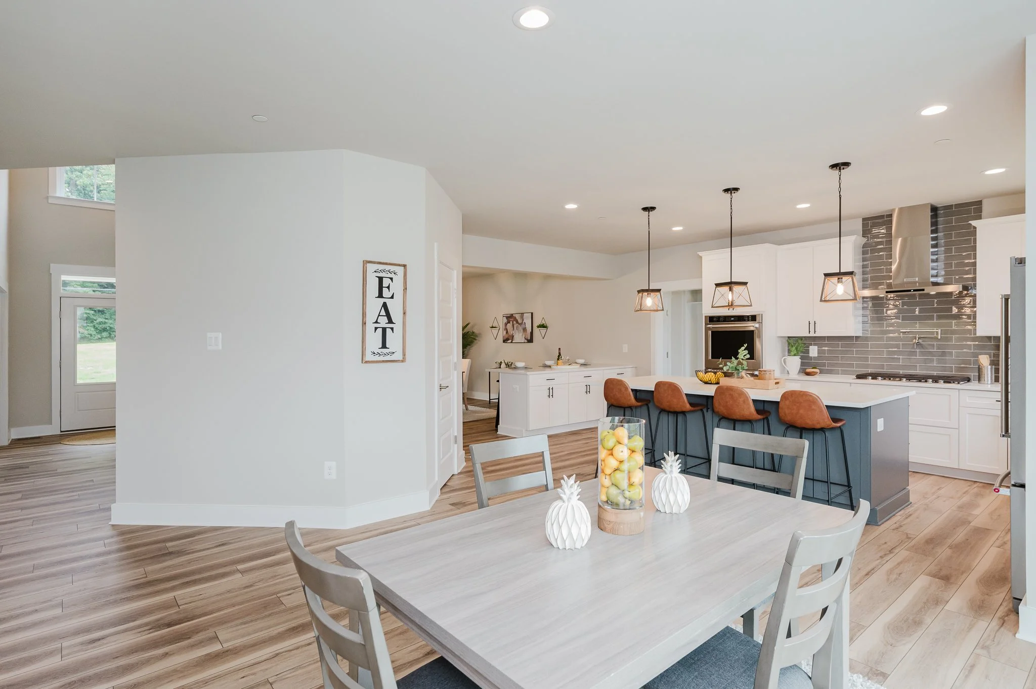 Open-concept kitchen and dining area with a light wood floor, white cabinets, a gray kitchen island with brown chairs, light fixtures, and a decorated dining table with pineapple-shaped decor and a fruit bowl.