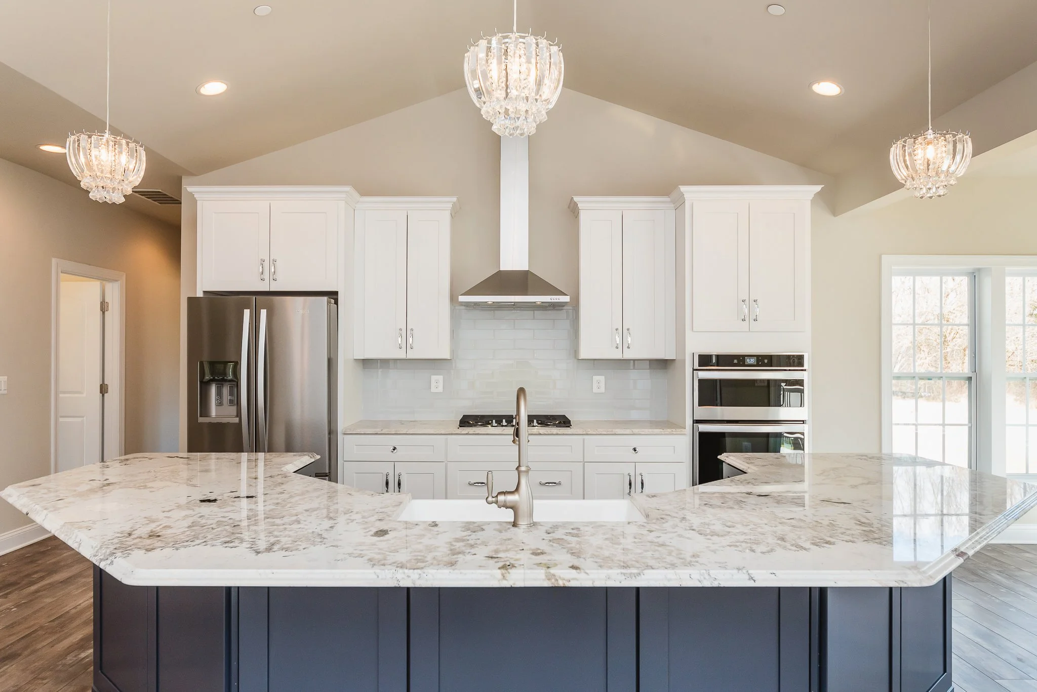Modern kitchen with white cabinetry, stainless steel refrigerator and oven, marble island with navy base, and three chandeliers hanging from the ceiling.