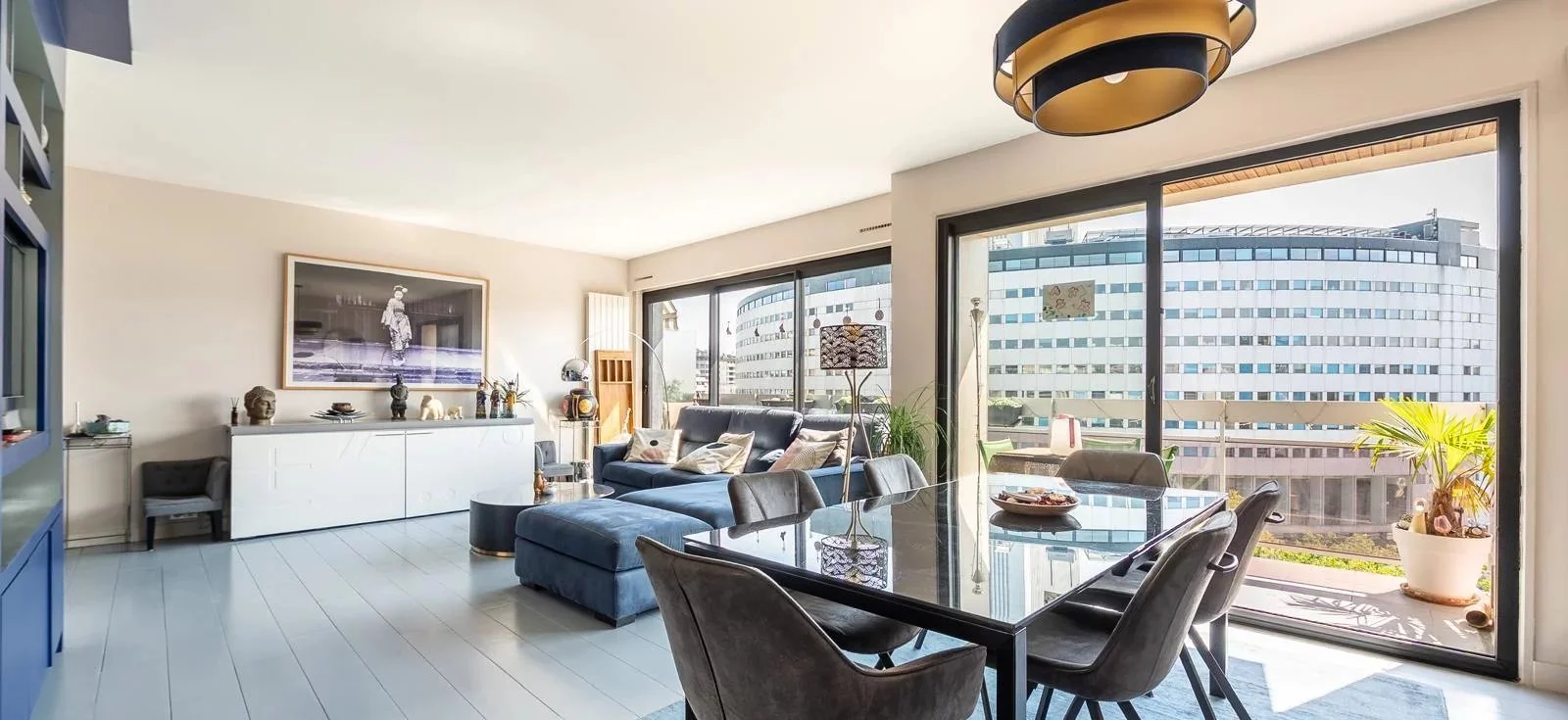 Salon moderne avec canapé bleu, table à manger noire, grandes fenêtres avec vue sur un immeuble de bureaux, et décoration intérieure élégante.